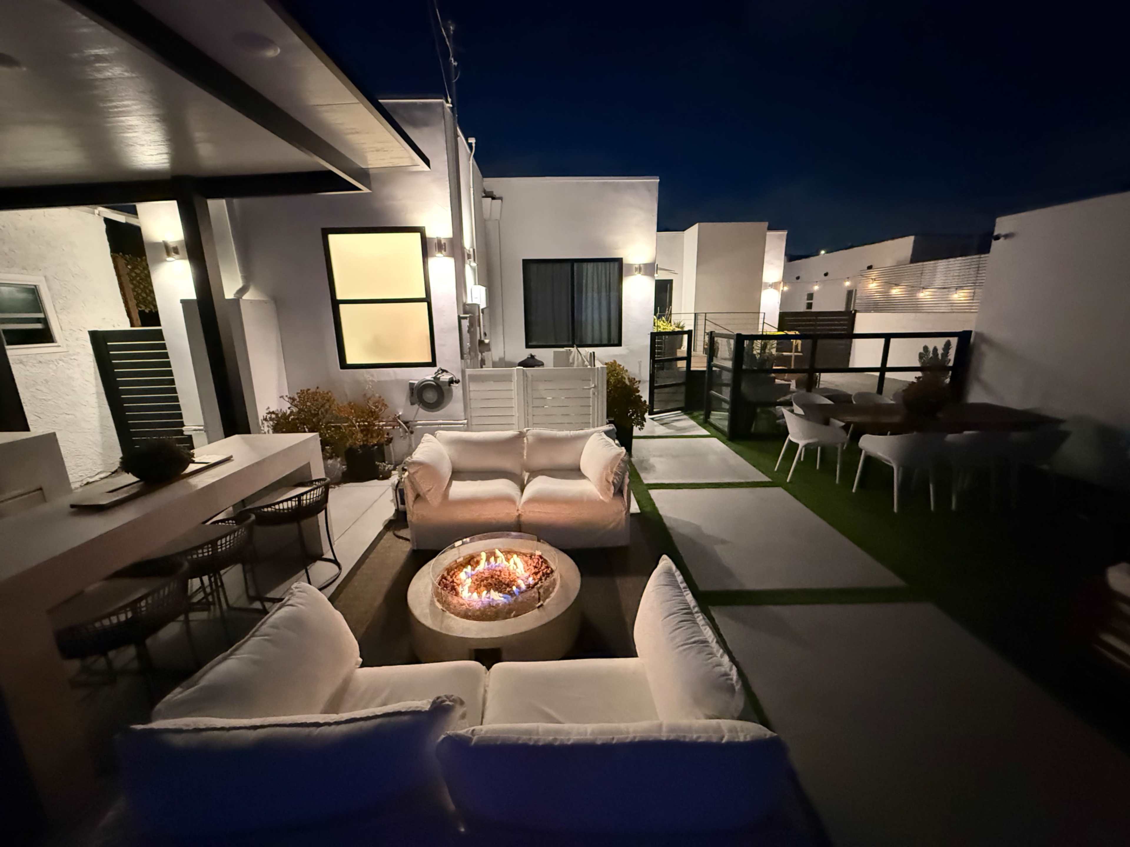 A modern outdoor seating area features a fire pit surrounded by sofas and a dining table, set against a backdrop of stylish architecture at night.
