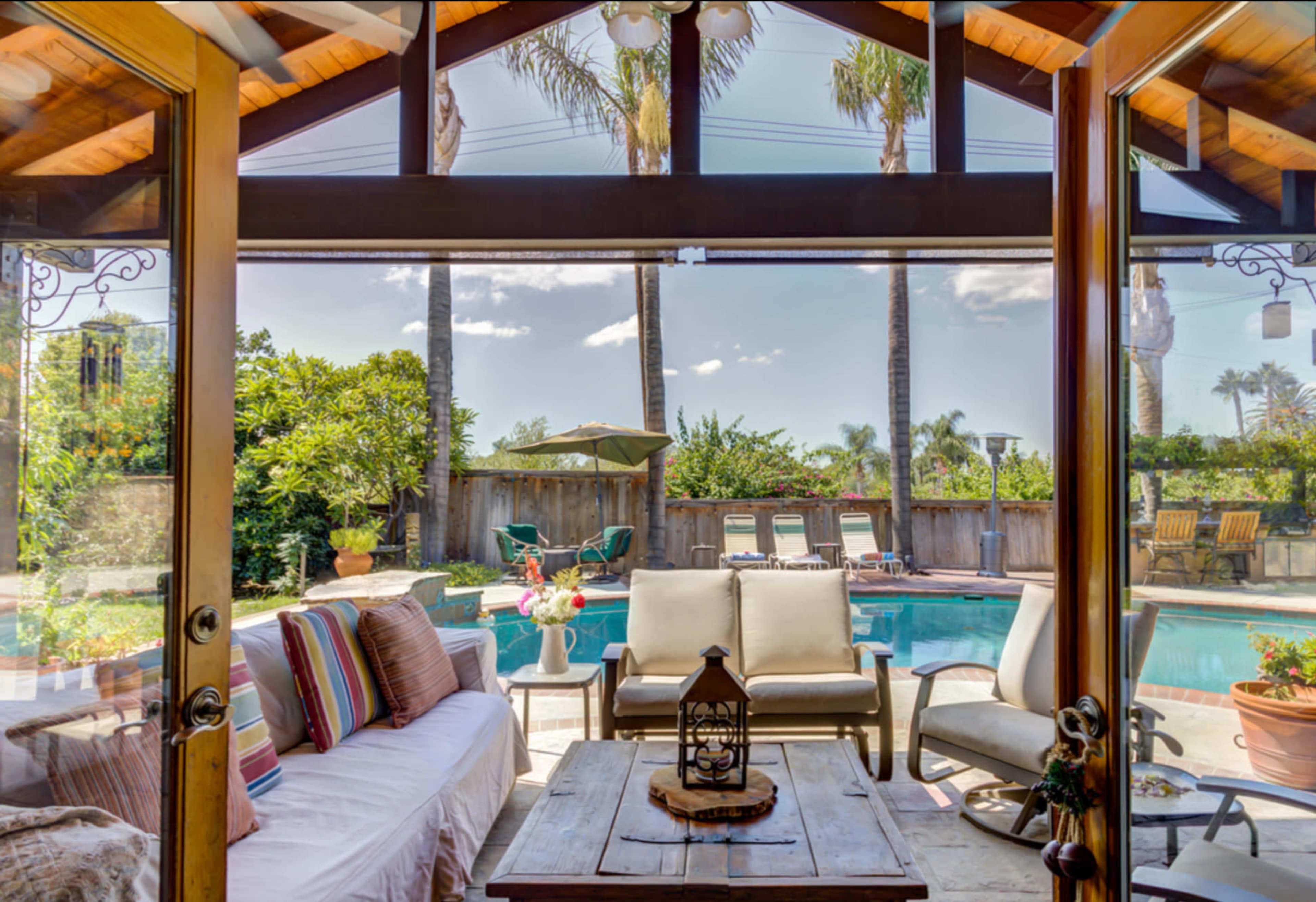 A view from inside a house through large glass doors, revealing a patio area with a seating arrangement and a swimming pool surrounded by palm trees.