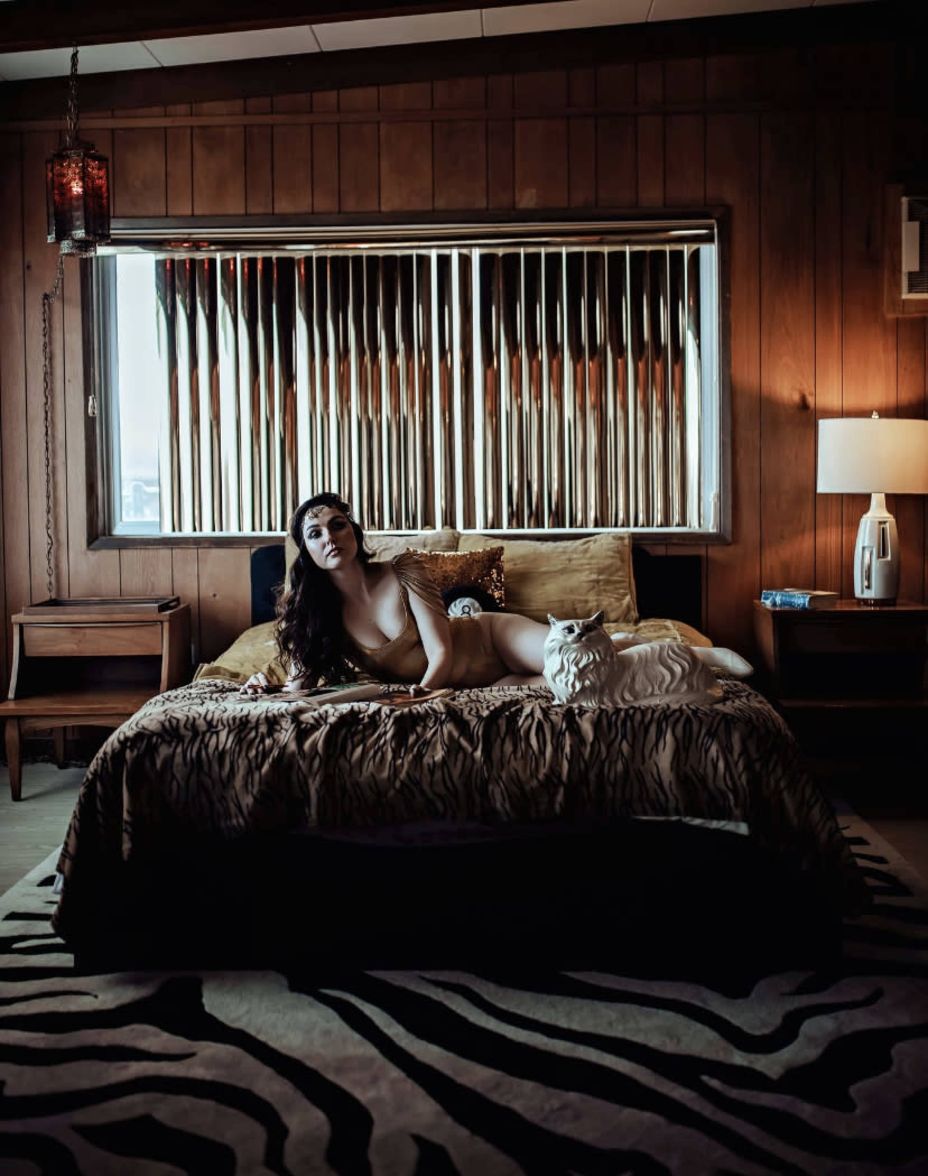 A woman lies on a bed with a tiger-striped cover, next to a white cat, in a room featuring wood-paneled walls and a window with vertical blinds.