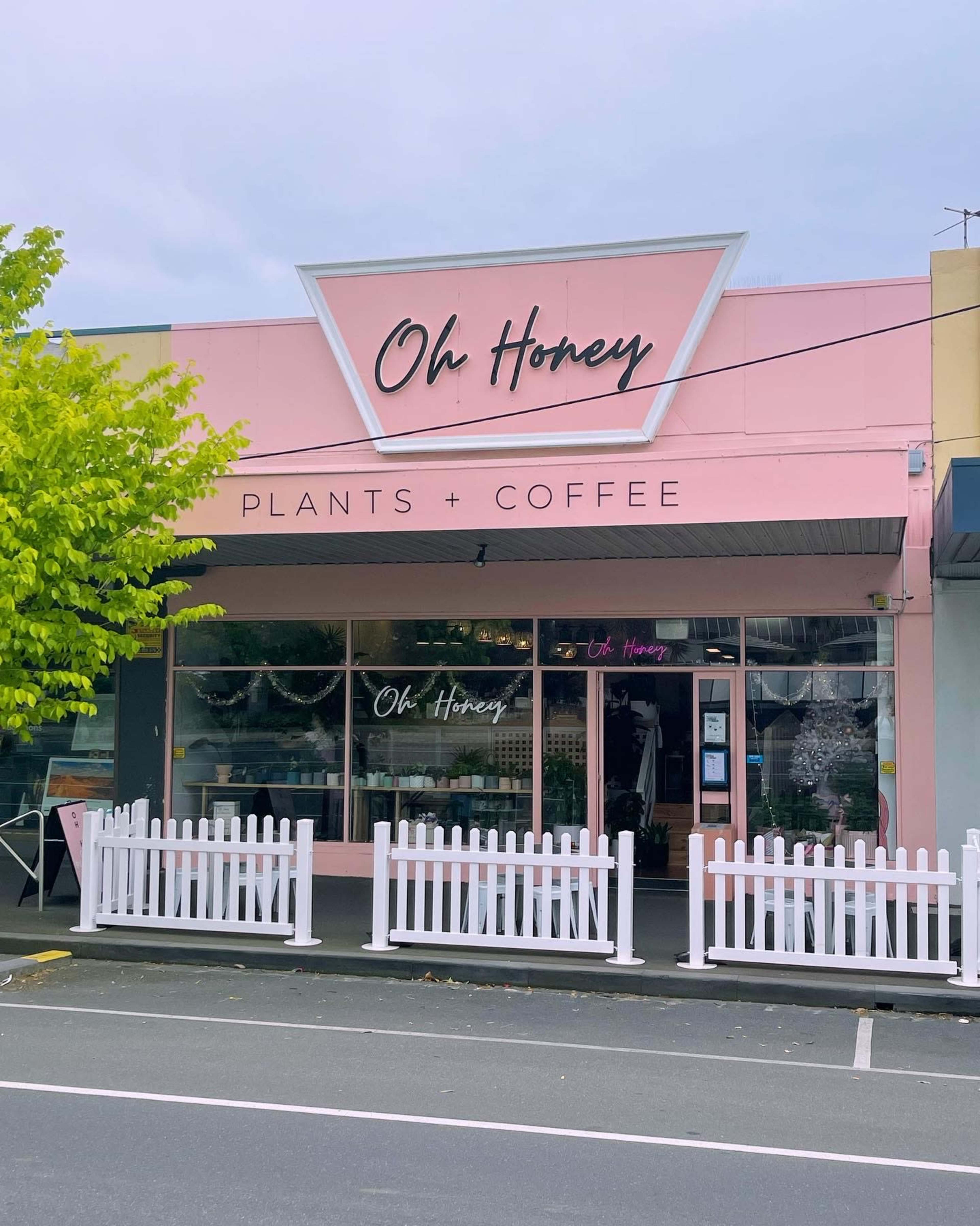 A pink storefront named "Oh Honey" that sells plants and coffee, featuring a white picket fence in front.