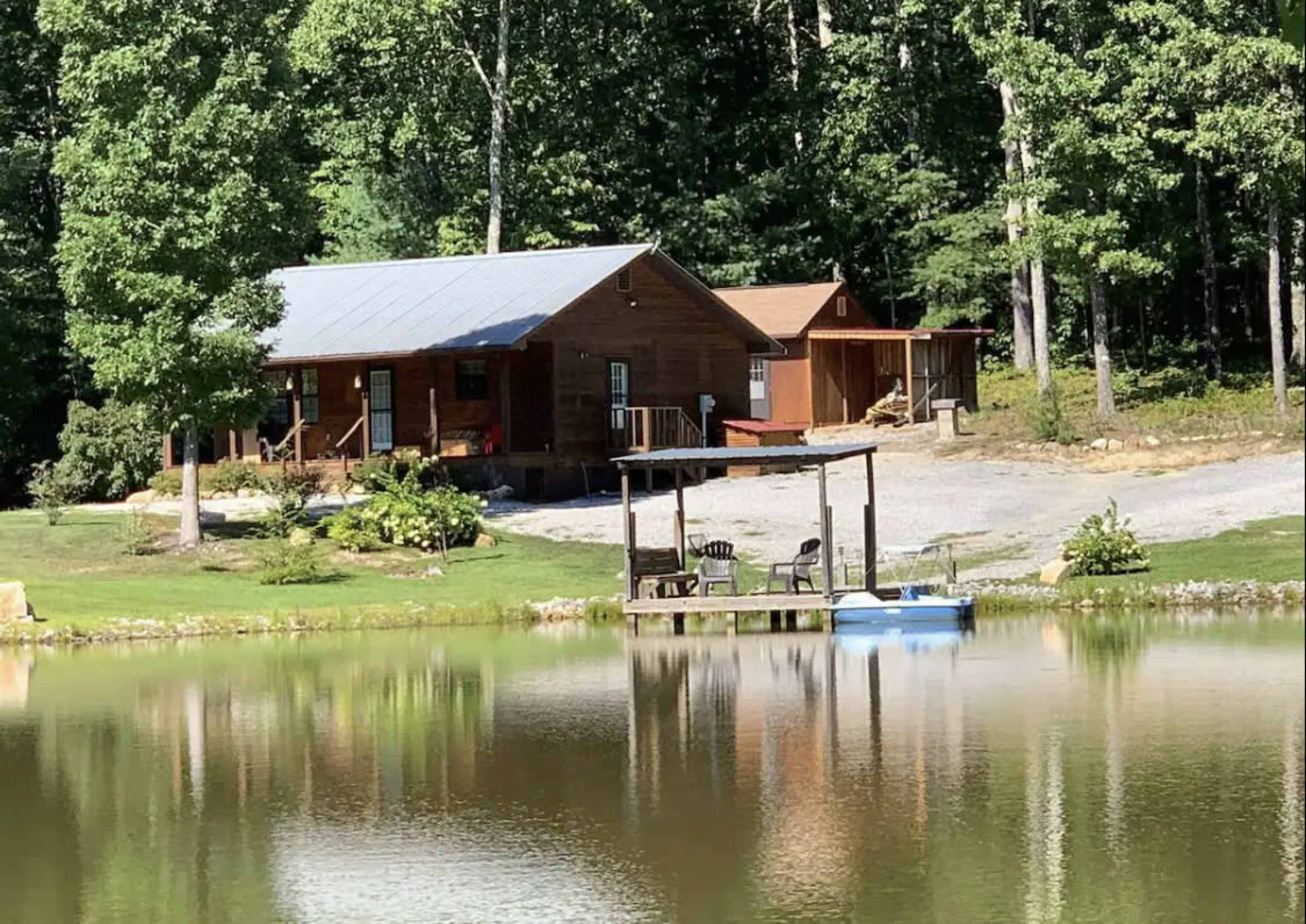 A wooden cabin with a metal roof is situated by a pond surrounded by trees.