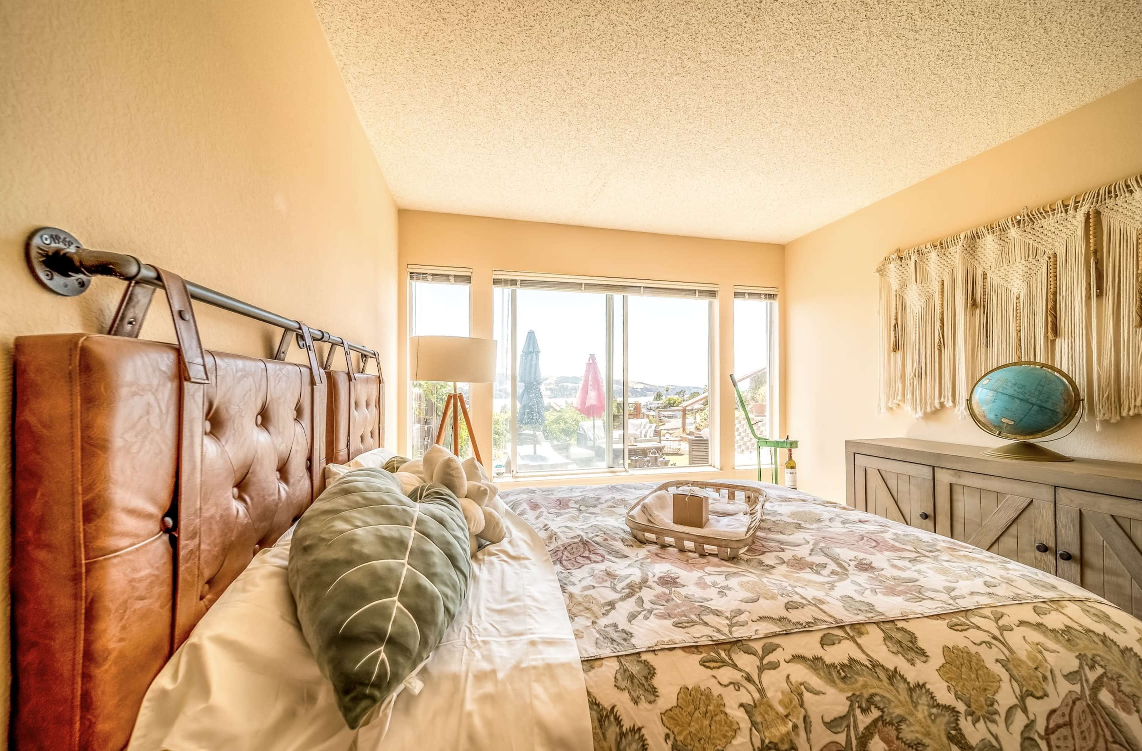 The image shows a cozy bedroom with a leather headboard, a floral bedspread, and large windows offering a view of the outdoors.
