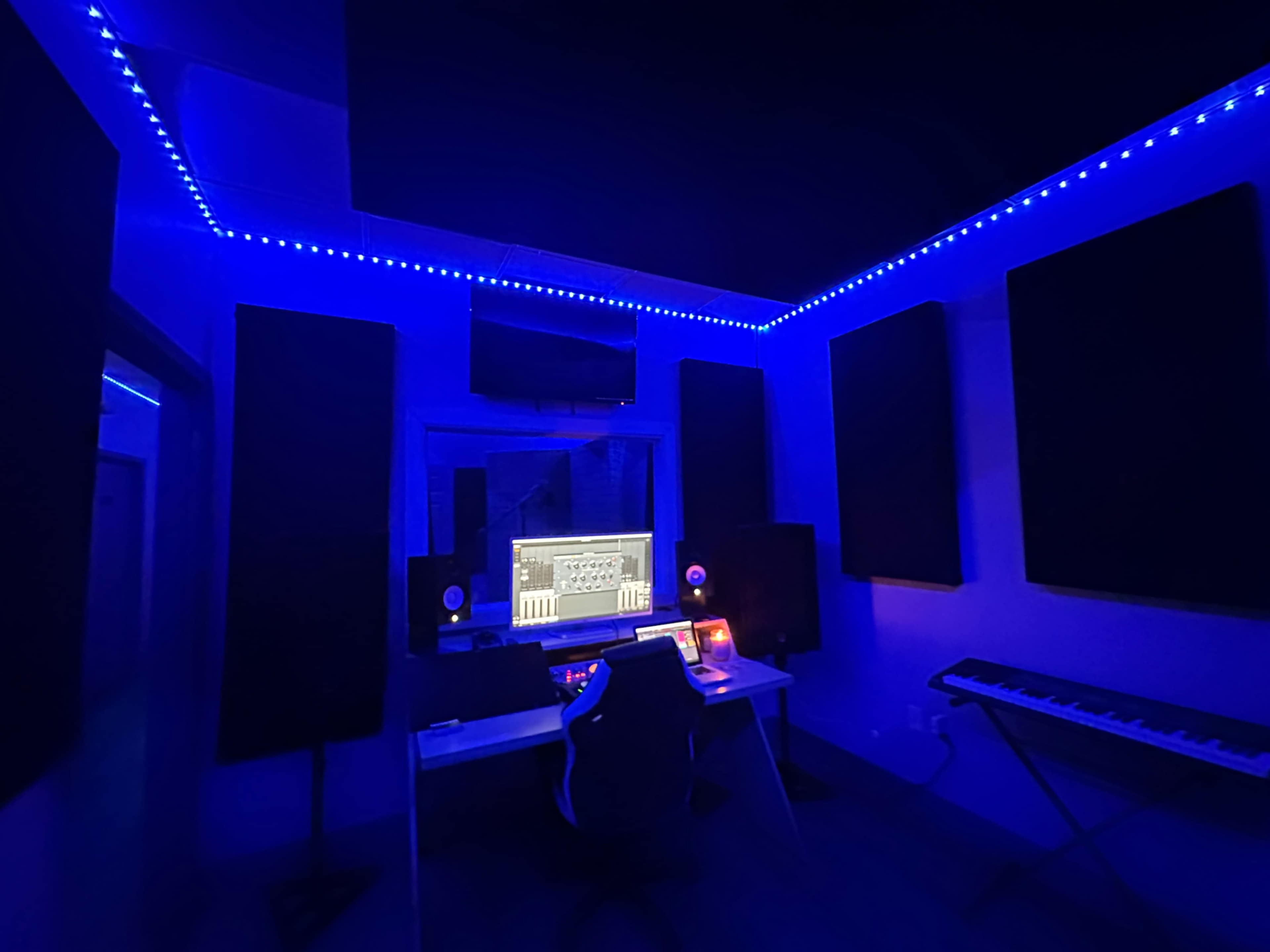 A dimly lit music studio with blue LED lights along the ceiling, a desk featuring a computer screen, speakers, and a keyboard.