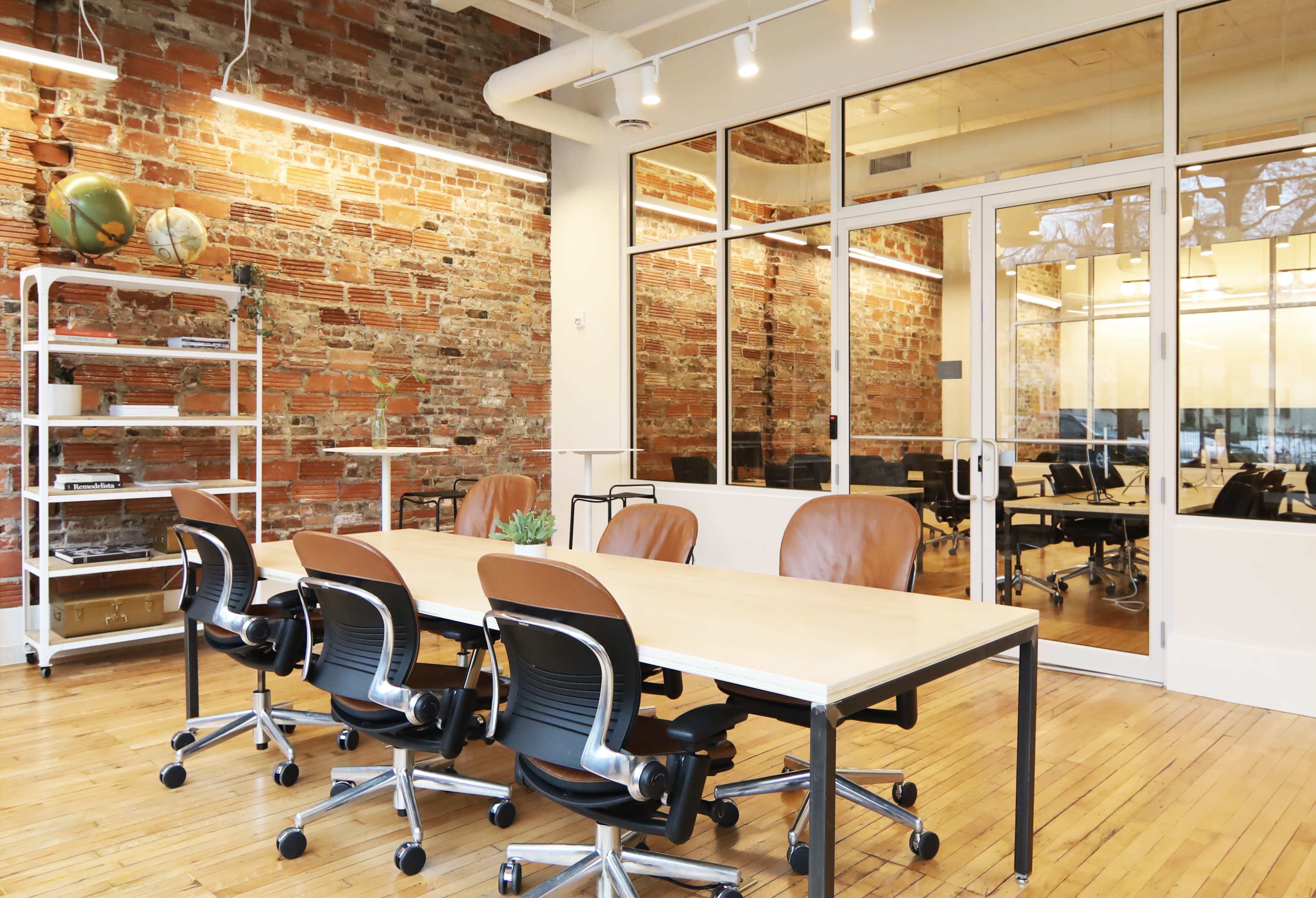 A modern office space with a long table and several ergonomic chairs, featuring exposed brick walls and large windows.