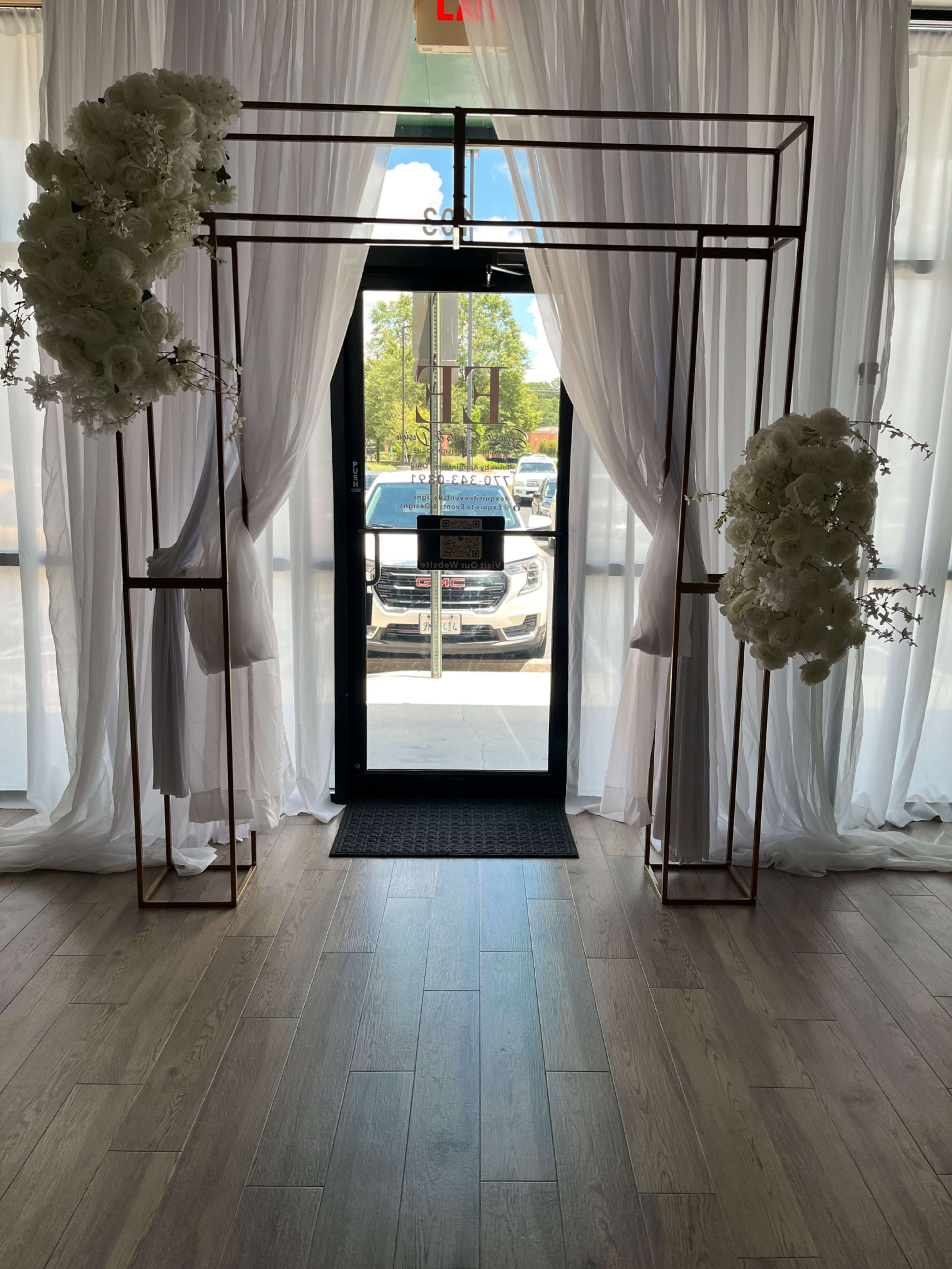 The image shows a decorative entrance arch made of metal with floral arrangements on either side, framed by sheer white curtains and leading to a doorway.