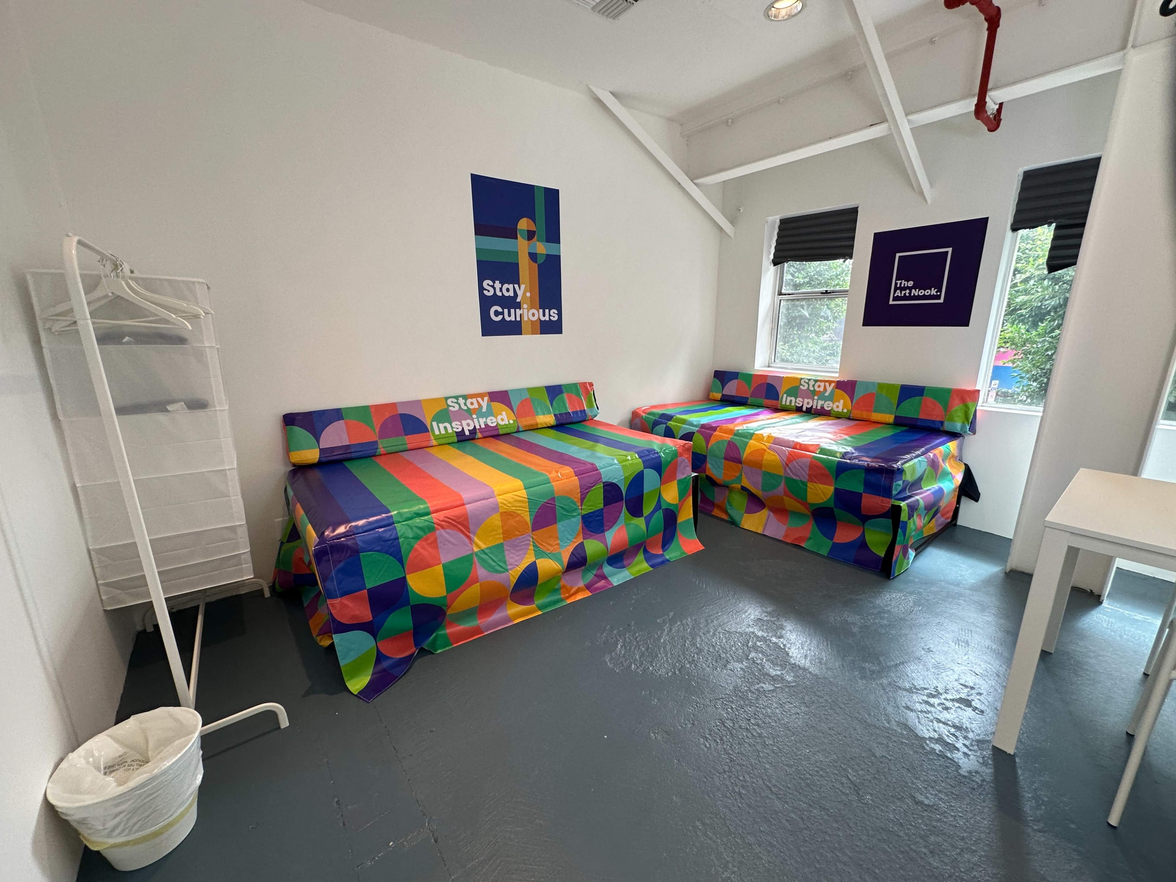 The image shows a brightly decorated room with two beds covered in colorful patterned bedspreads, a wardrobe, a waste bin, and posters on the walls.