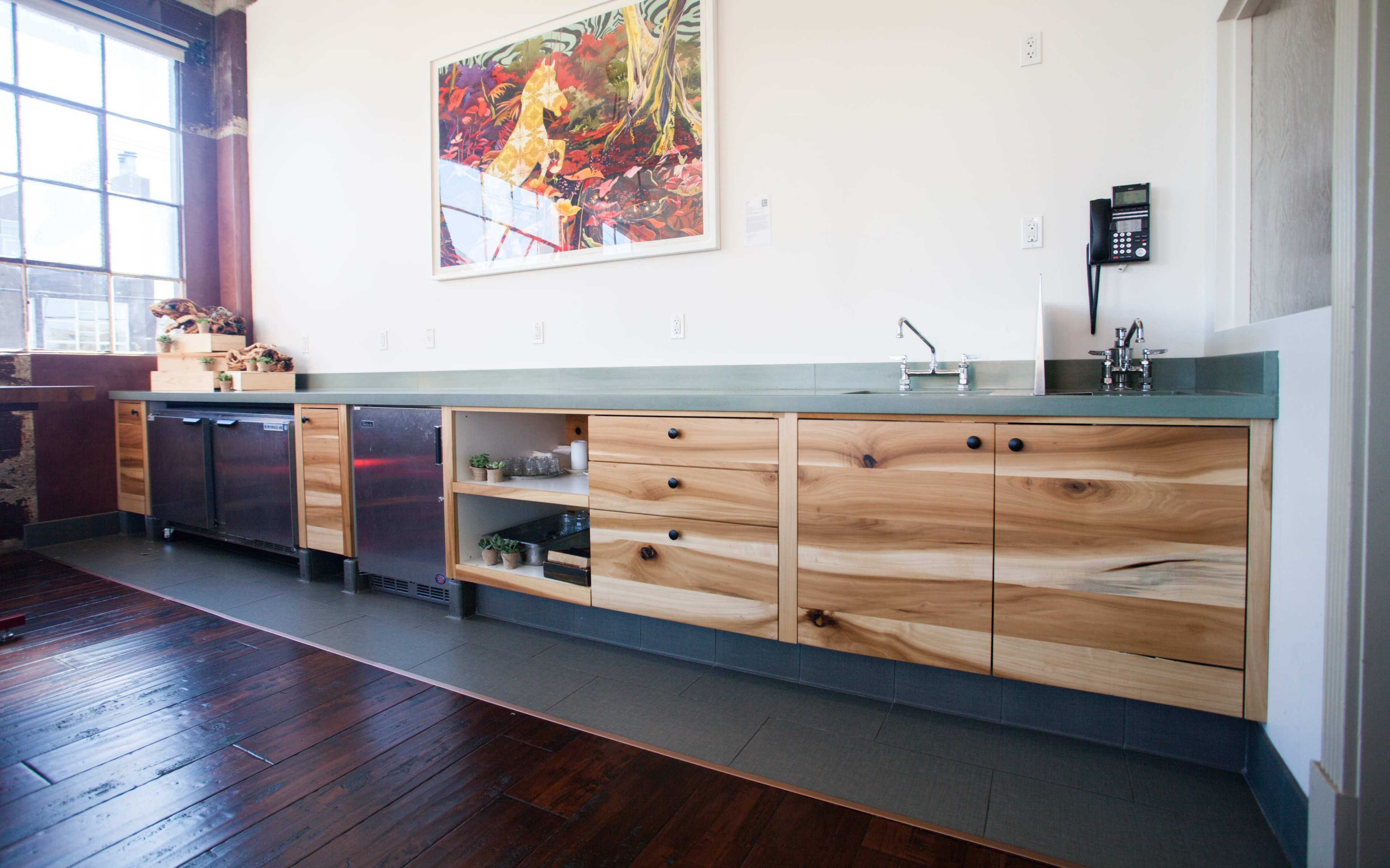 A modern kitchen area with wooden cabinets, a stainless steel refrigerator, a sink, and a stone countertop under a large window.