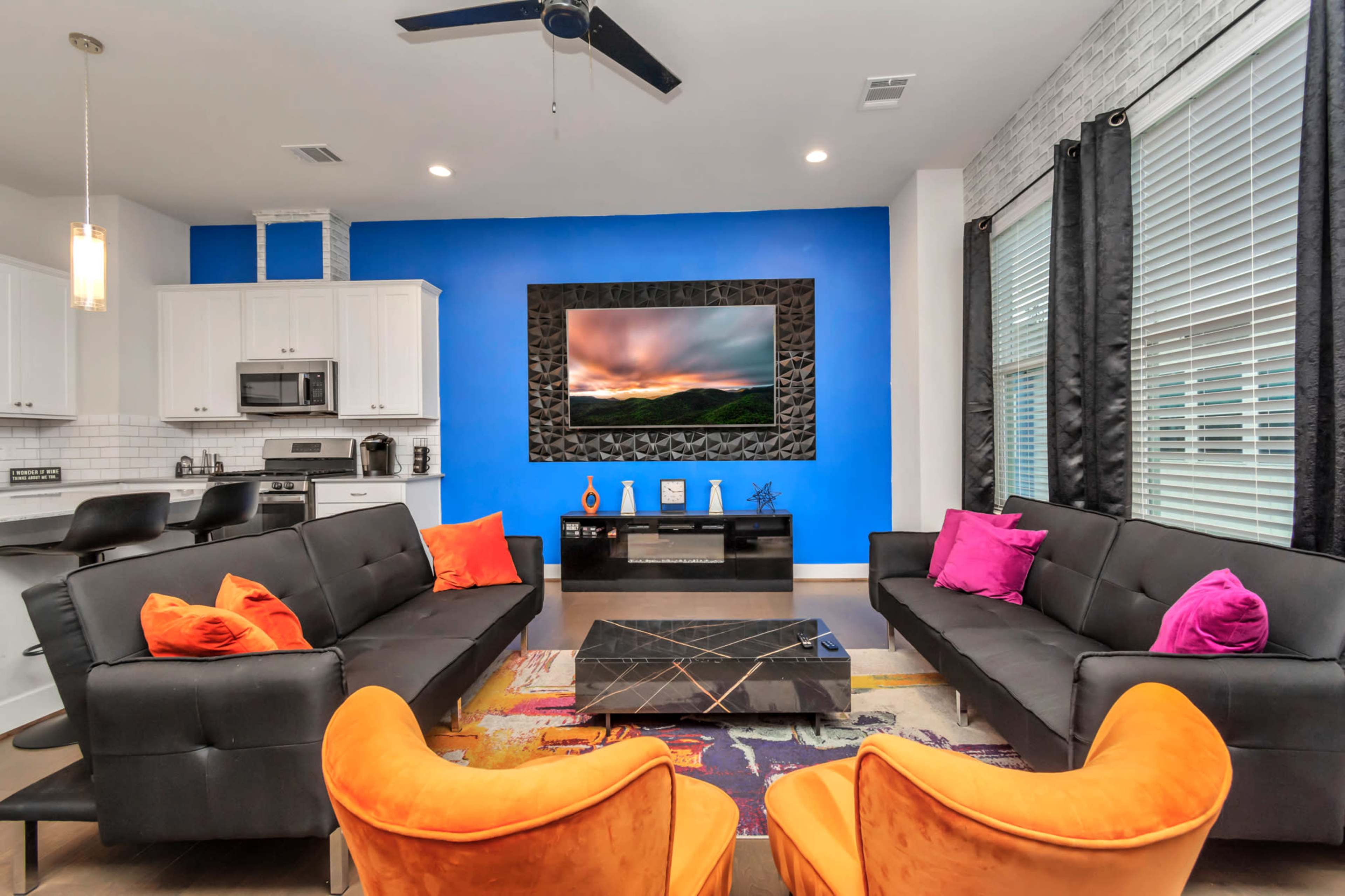 The room features a bright blue accent wall, a large flat-screen TV mounted above a stone frame, modern black sofas with colorful throw pillows, and an orange armchair, all set against a backdrop of large windows.