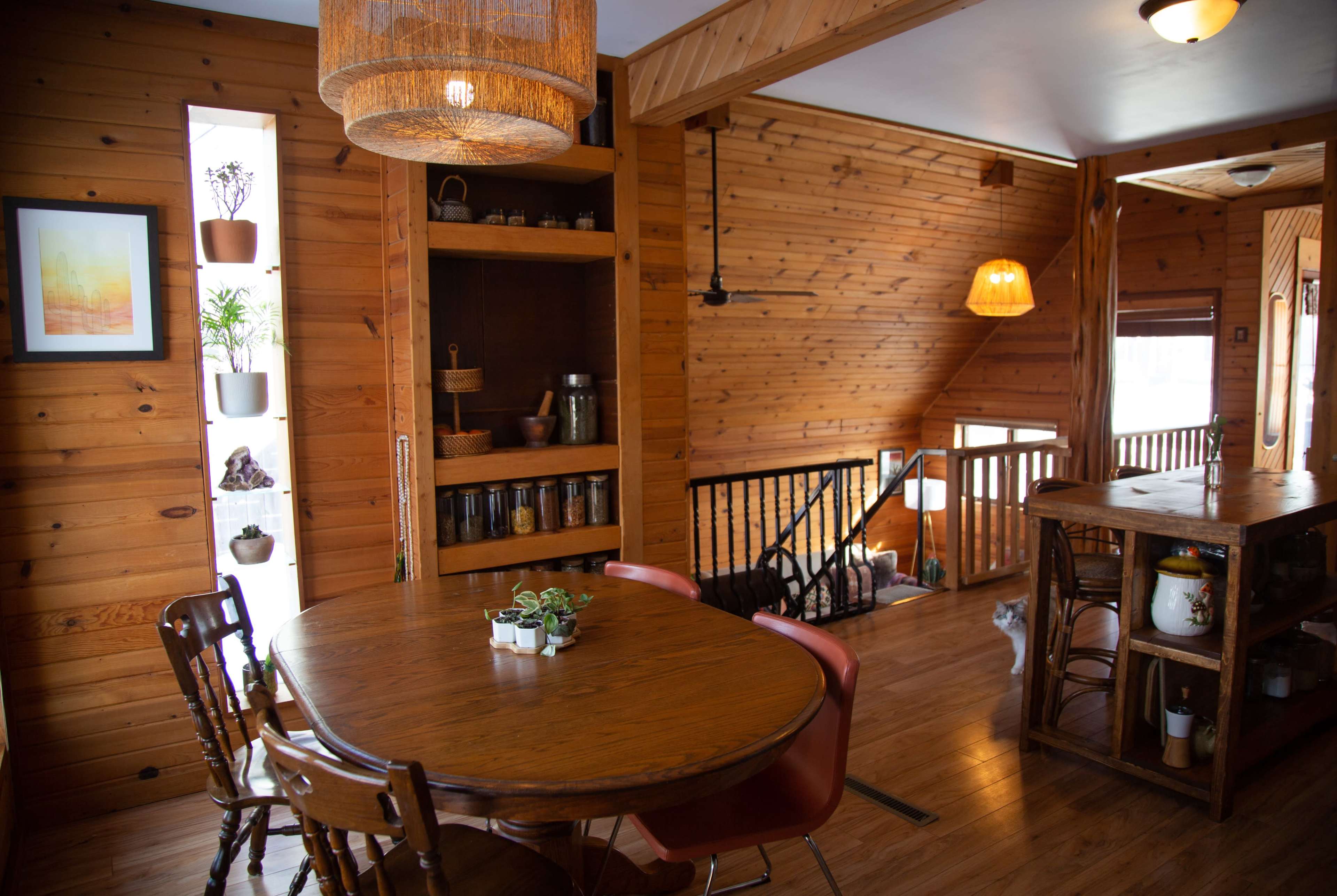 A wooden interior of a home featuring a dining table with chairs, a bookshelf, and an open staircase.