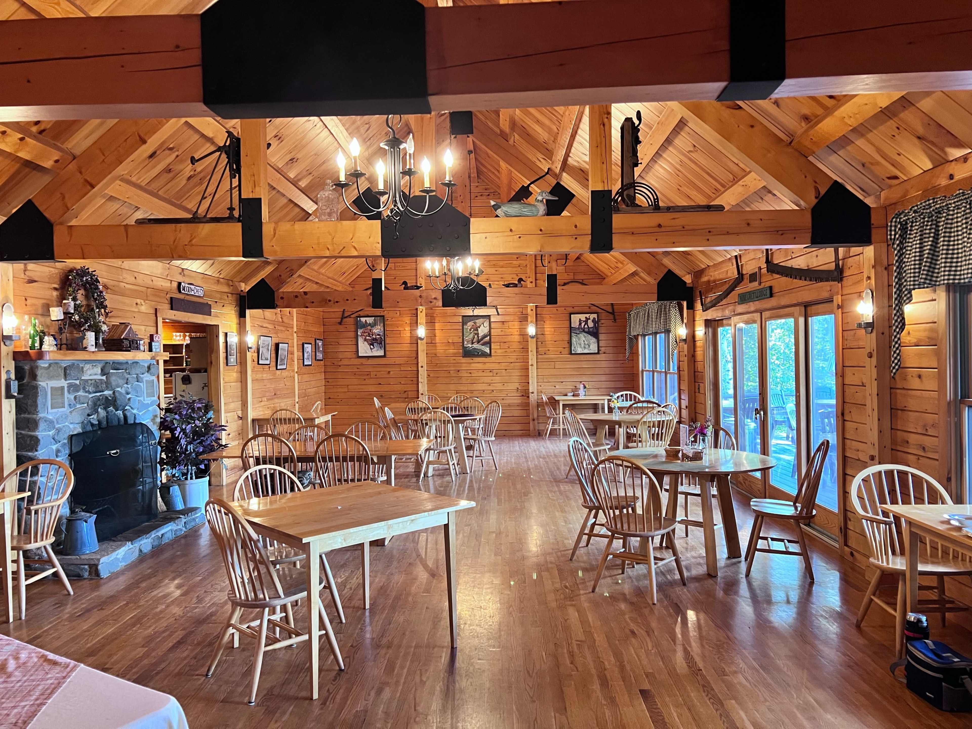The image shows a wooden dining room with tables and chairs arranged around a stone fireplace, illuminated by hanging lights.
