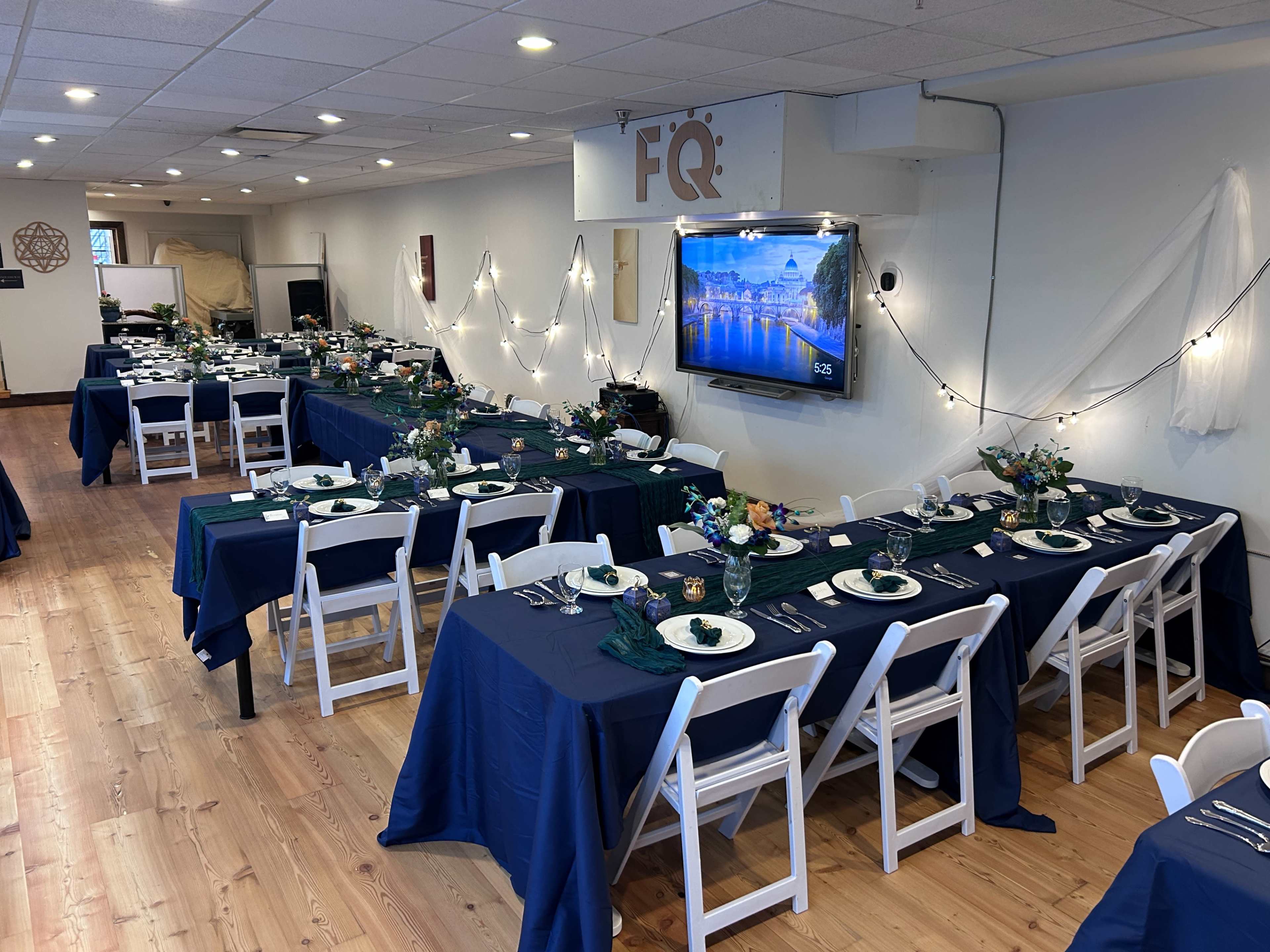 The image shows a dining setup with multiple tables covered in navy blue tablecloths, arranged in a well-lit indoor space adorned with decorations and lighting.