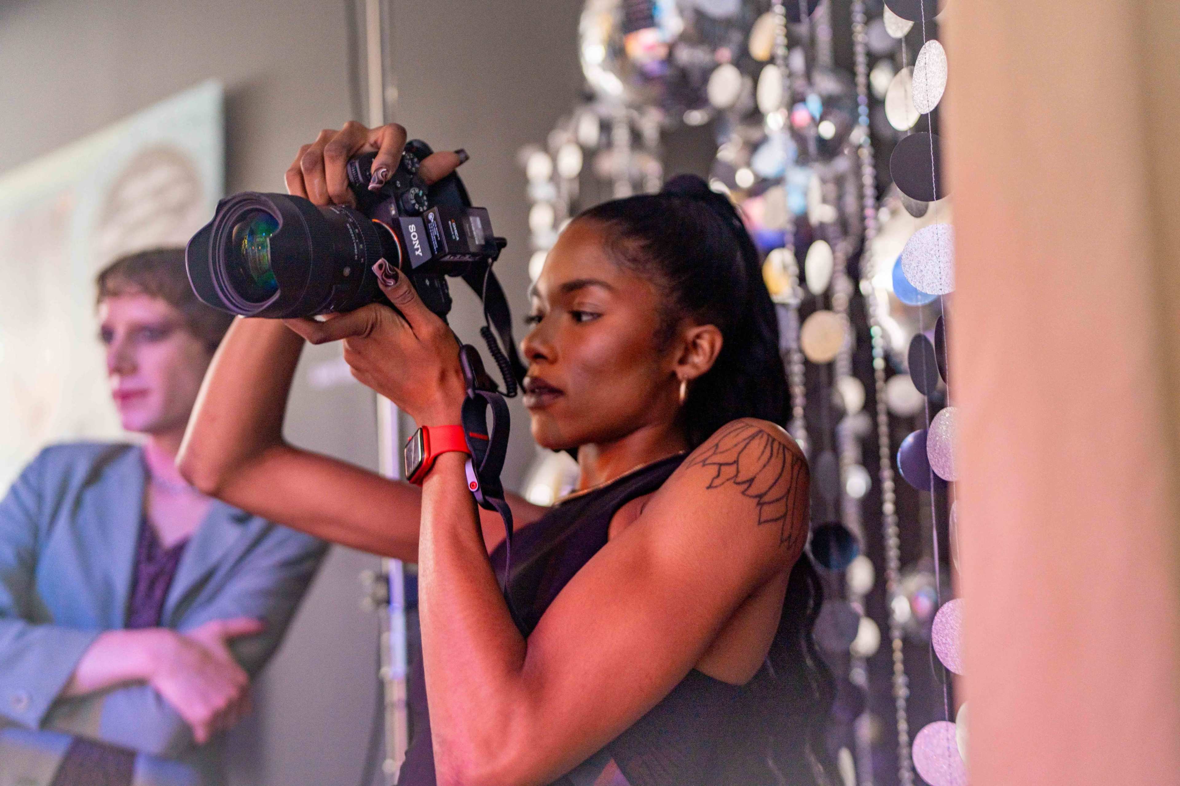 A woman with a camera is taking a photograph in front of a decorated background, while another person stands nearby observing.