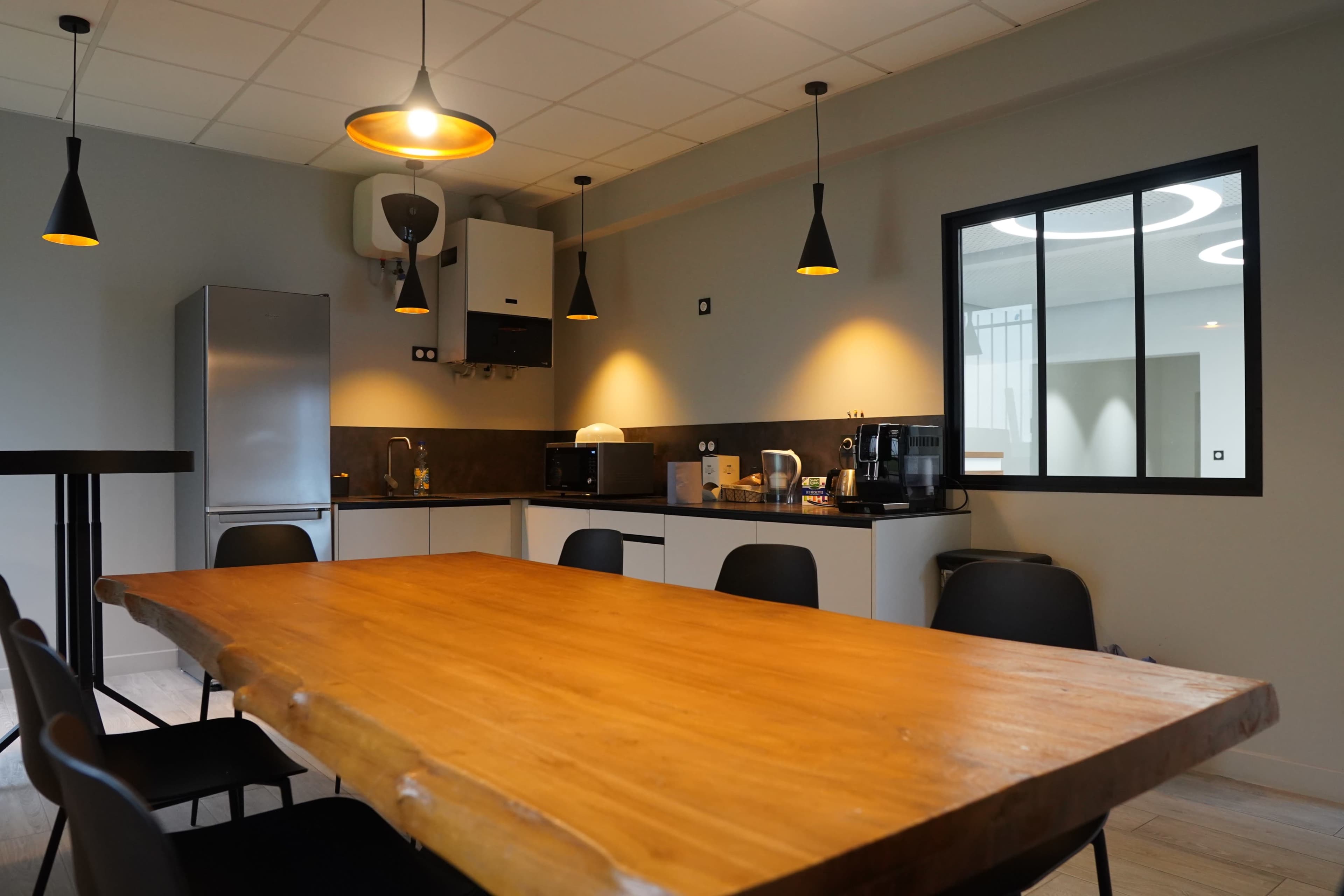 A modern kitchen with a large wooden table, black chairs, and various appliances including a fridge and coffee maker.
