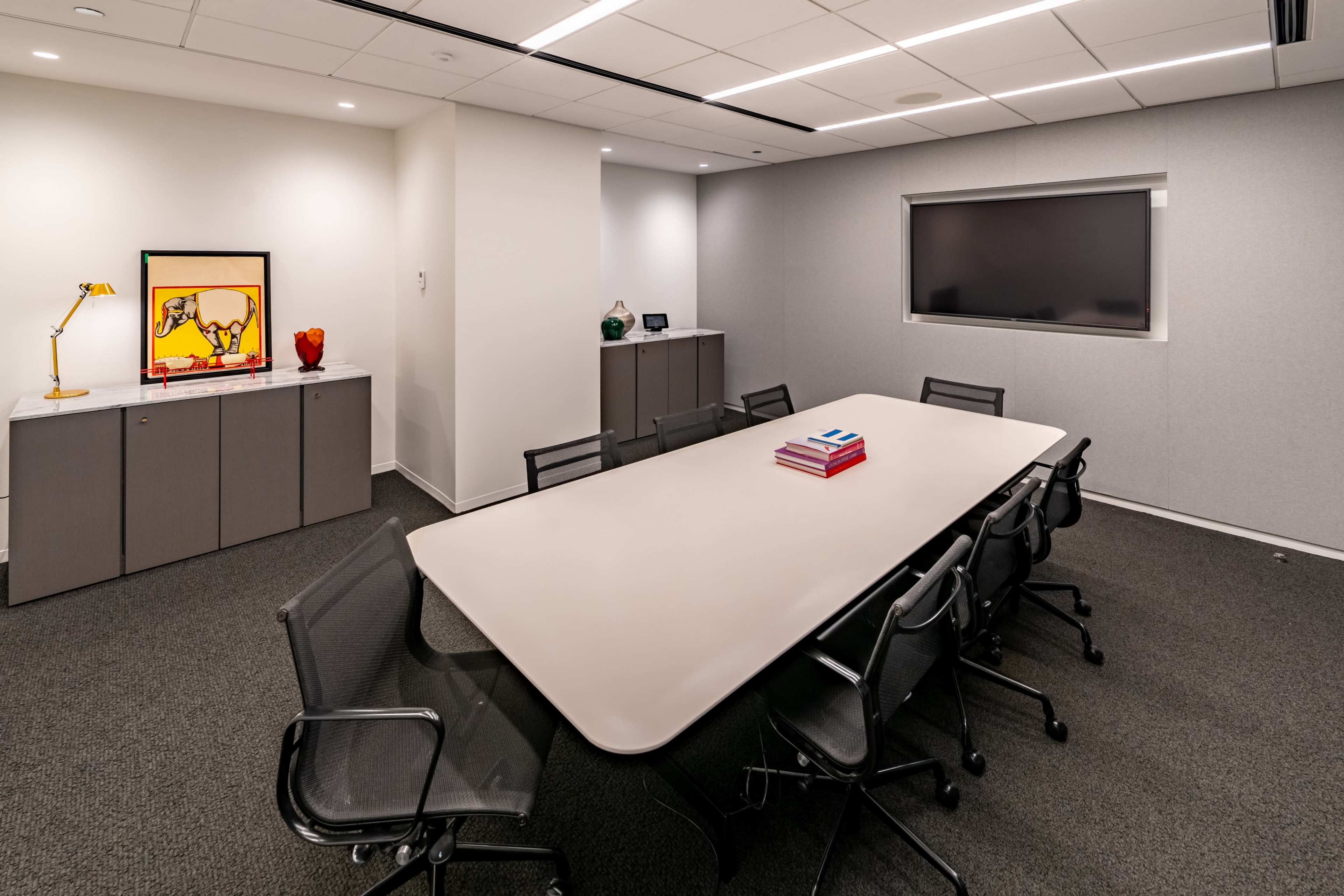 A modern conference room features a long table surrounded by black chairs, with a colorful artwork displayed on the wall and multiple cabinets along the sides.