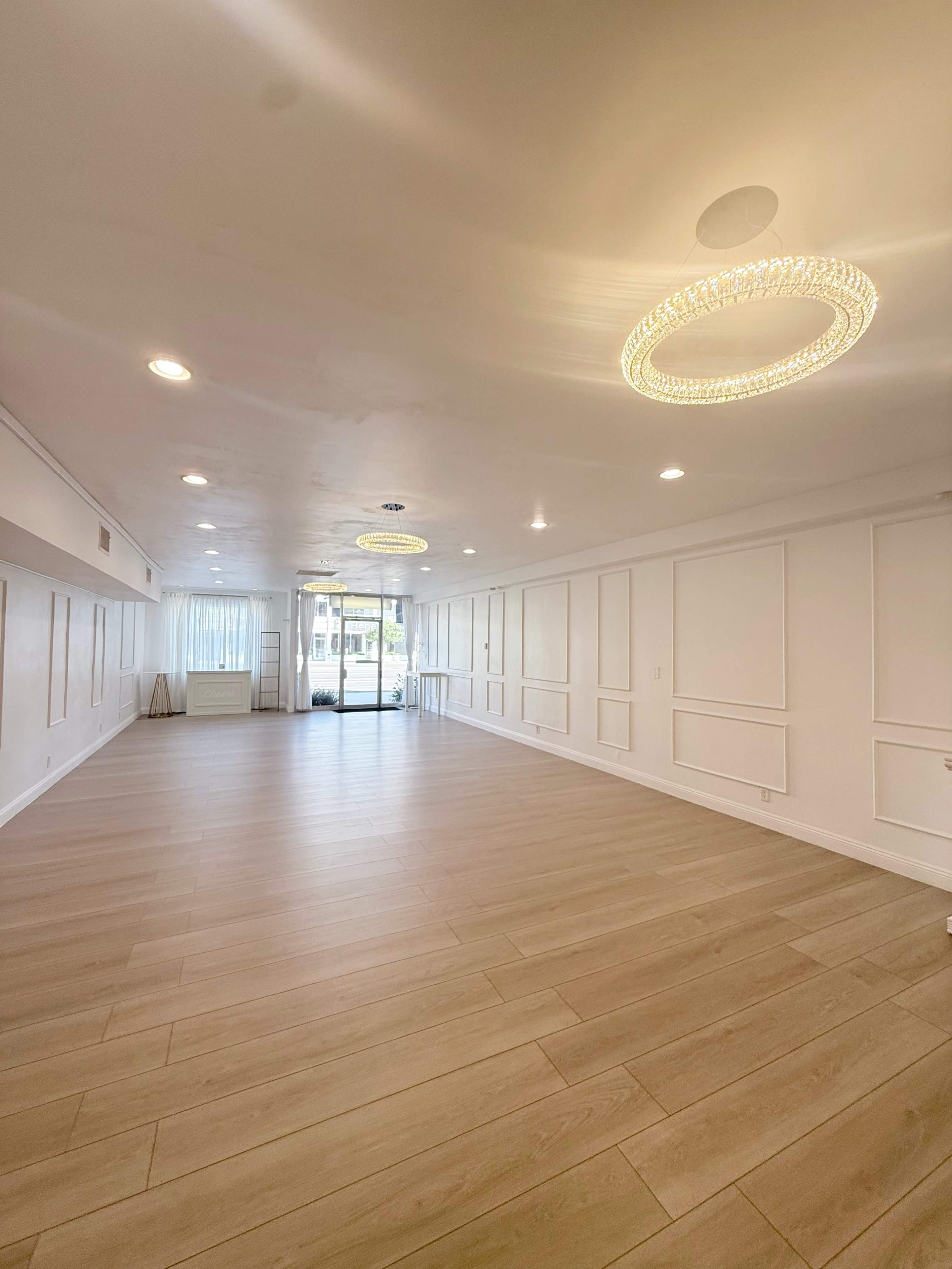 The image shows a spacious, well-lit room with wooden flooring and modern chandeliers, featuring white walls and large windows at one end.