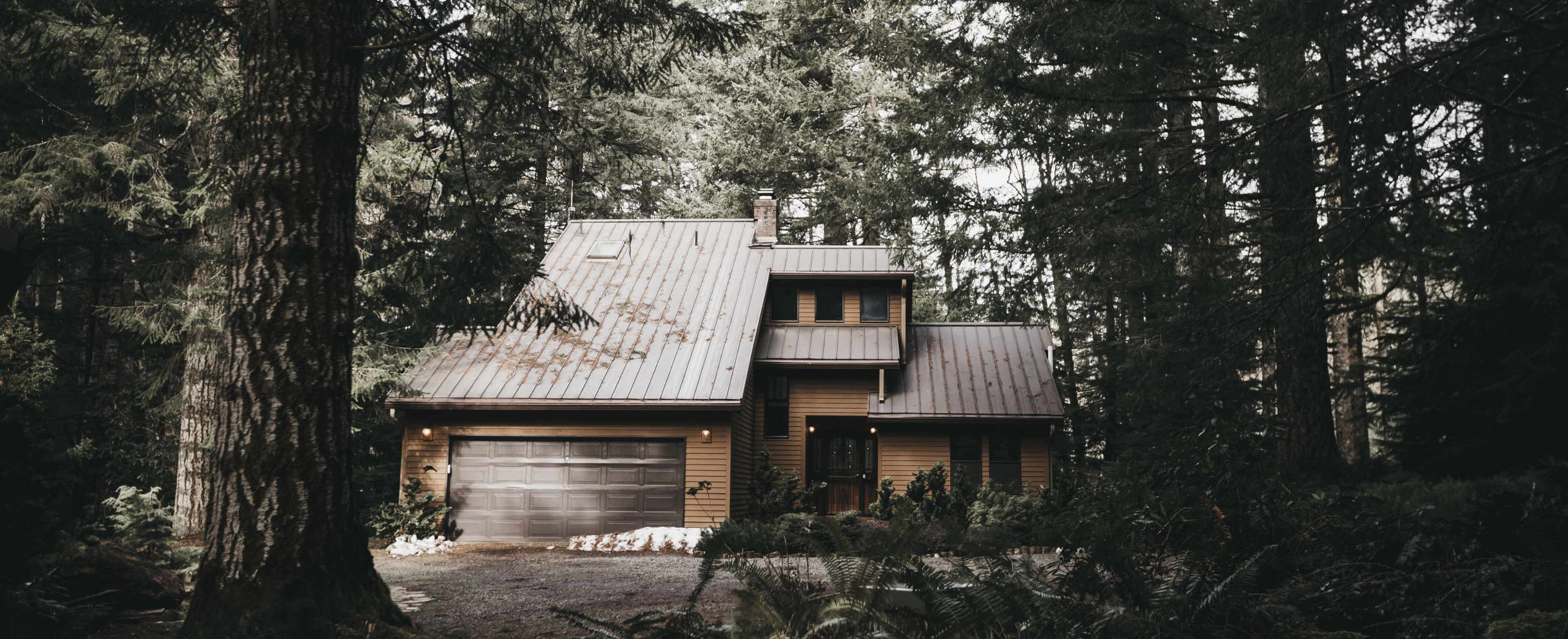 A two-story wooden house with a metal roof is situated among tall trees in a forested area.