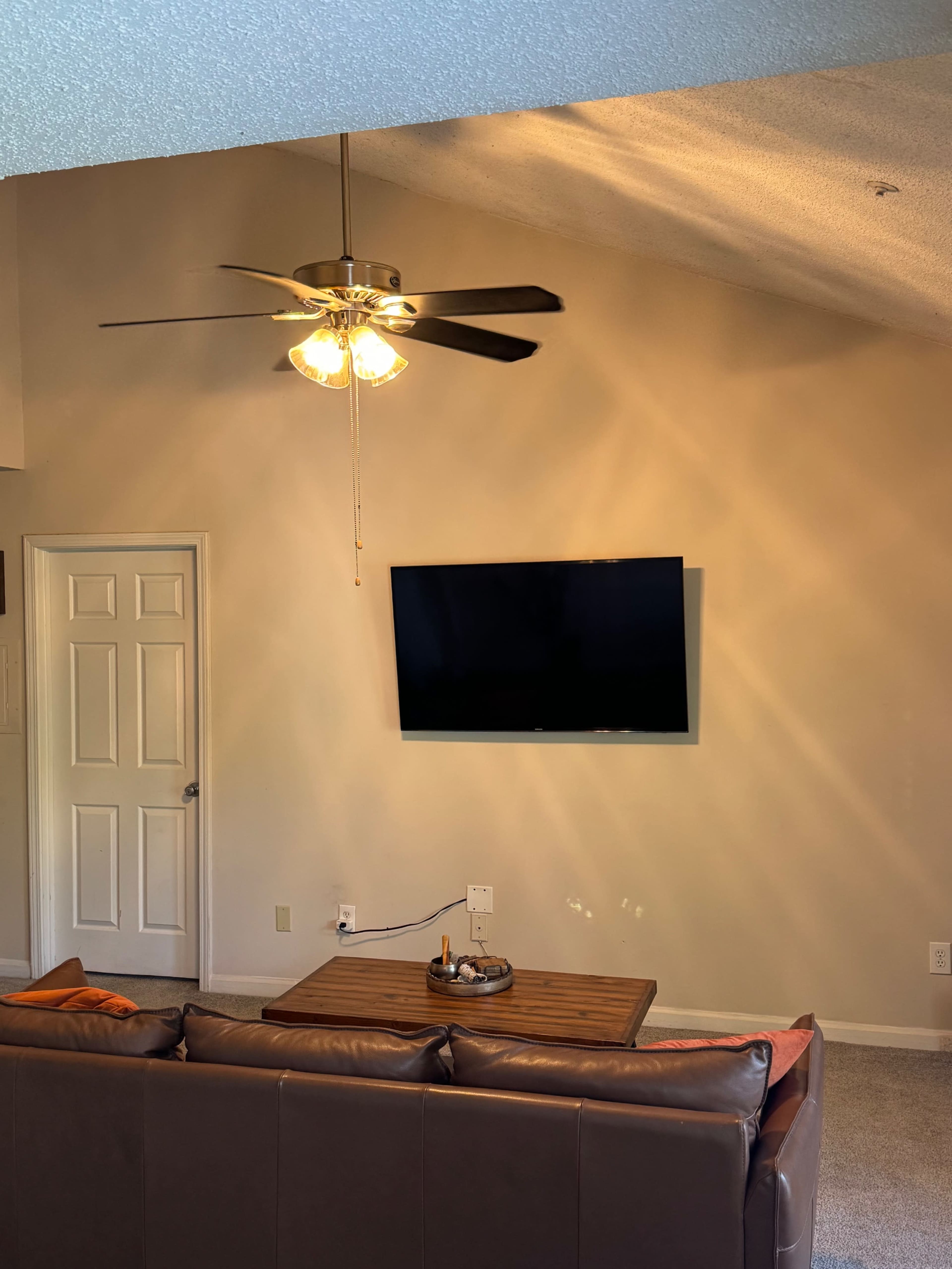 The image displays a living room with a leather couch facing a wall-mounted television and a ceiling fan above.