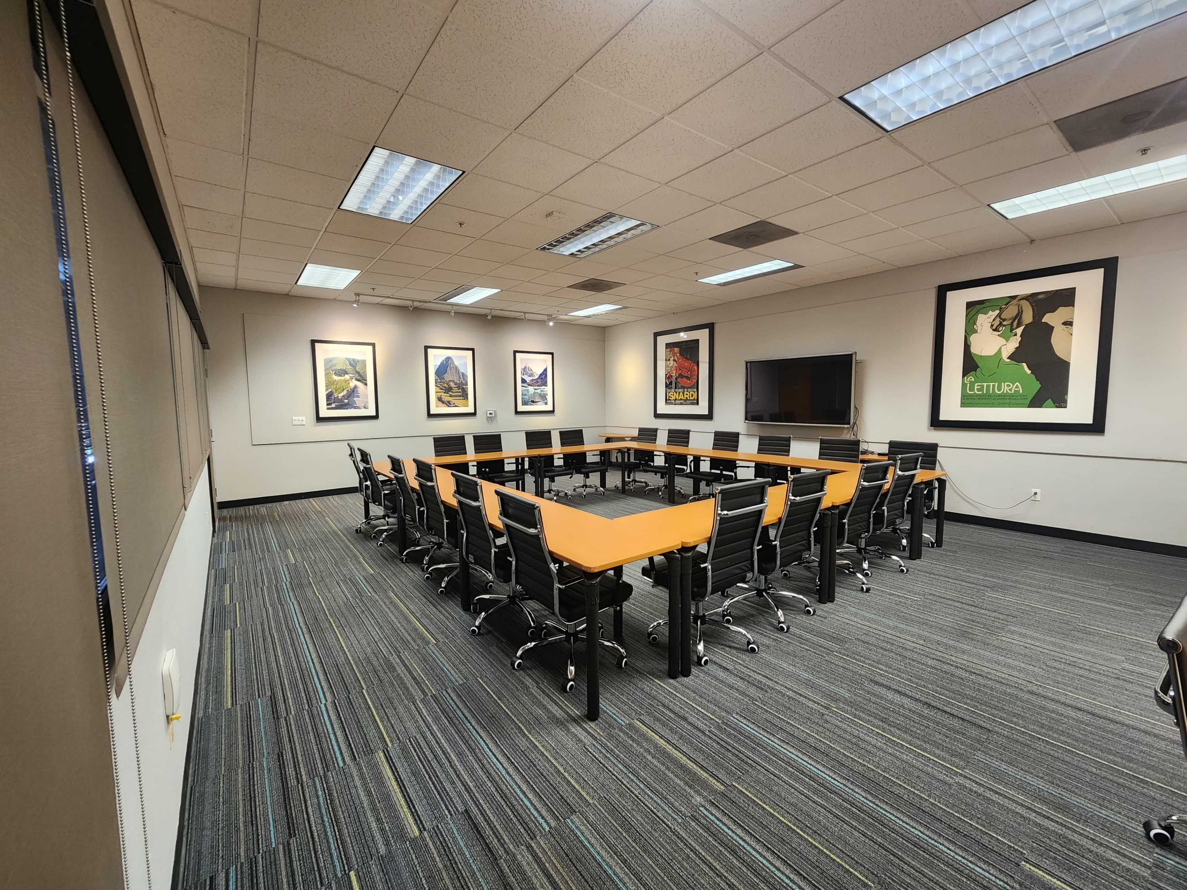 The image shows a modern conference room with a rectangular table surrounded by black chairs, featuring colorful framed artwork on the walls and a large screen at one end.