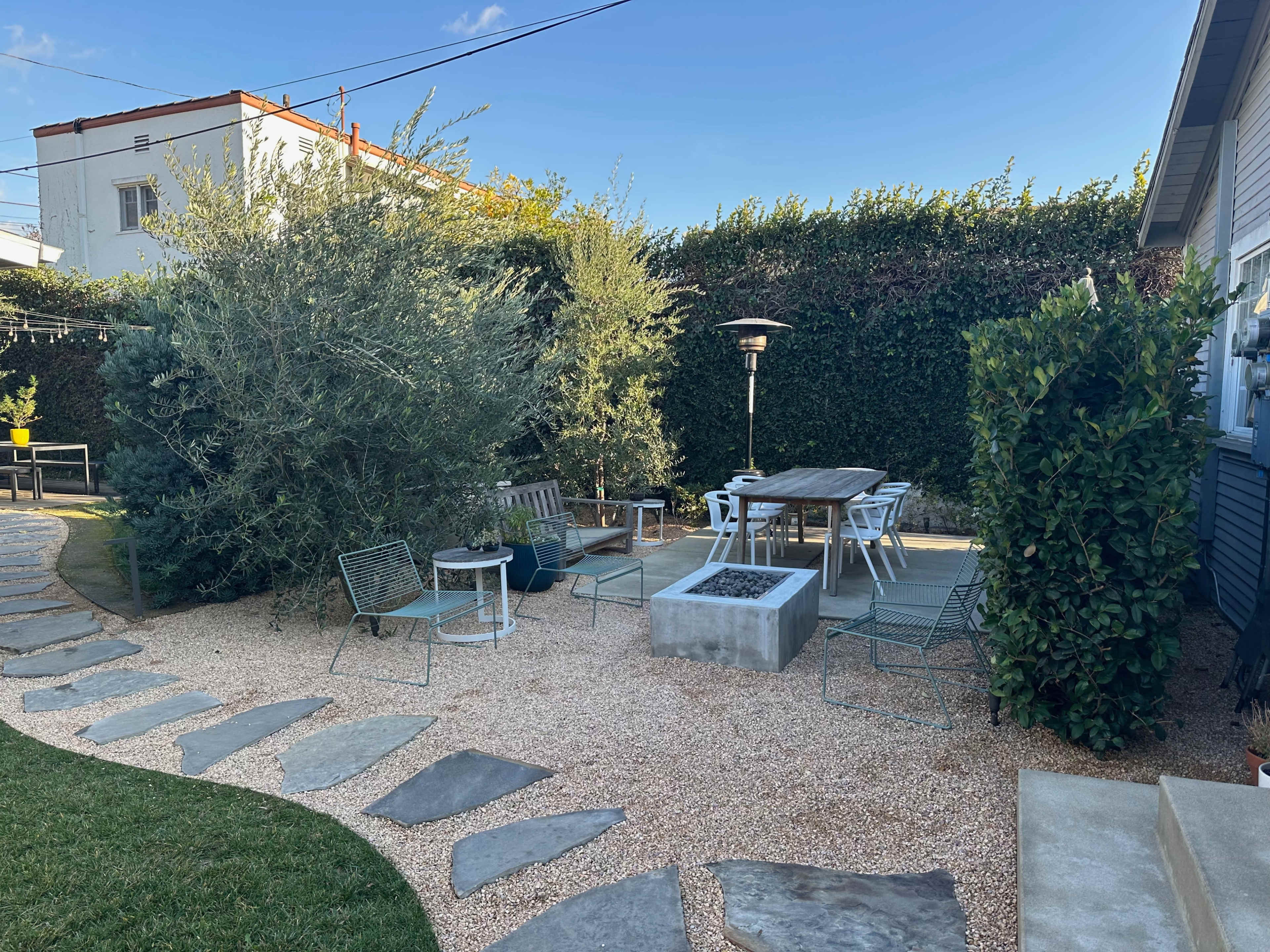 The image depicts a landscaped outdoor seating area featuring stone pathways, gravel ground, a wooden table surrounded by chairs, and greenery along the perimeter.