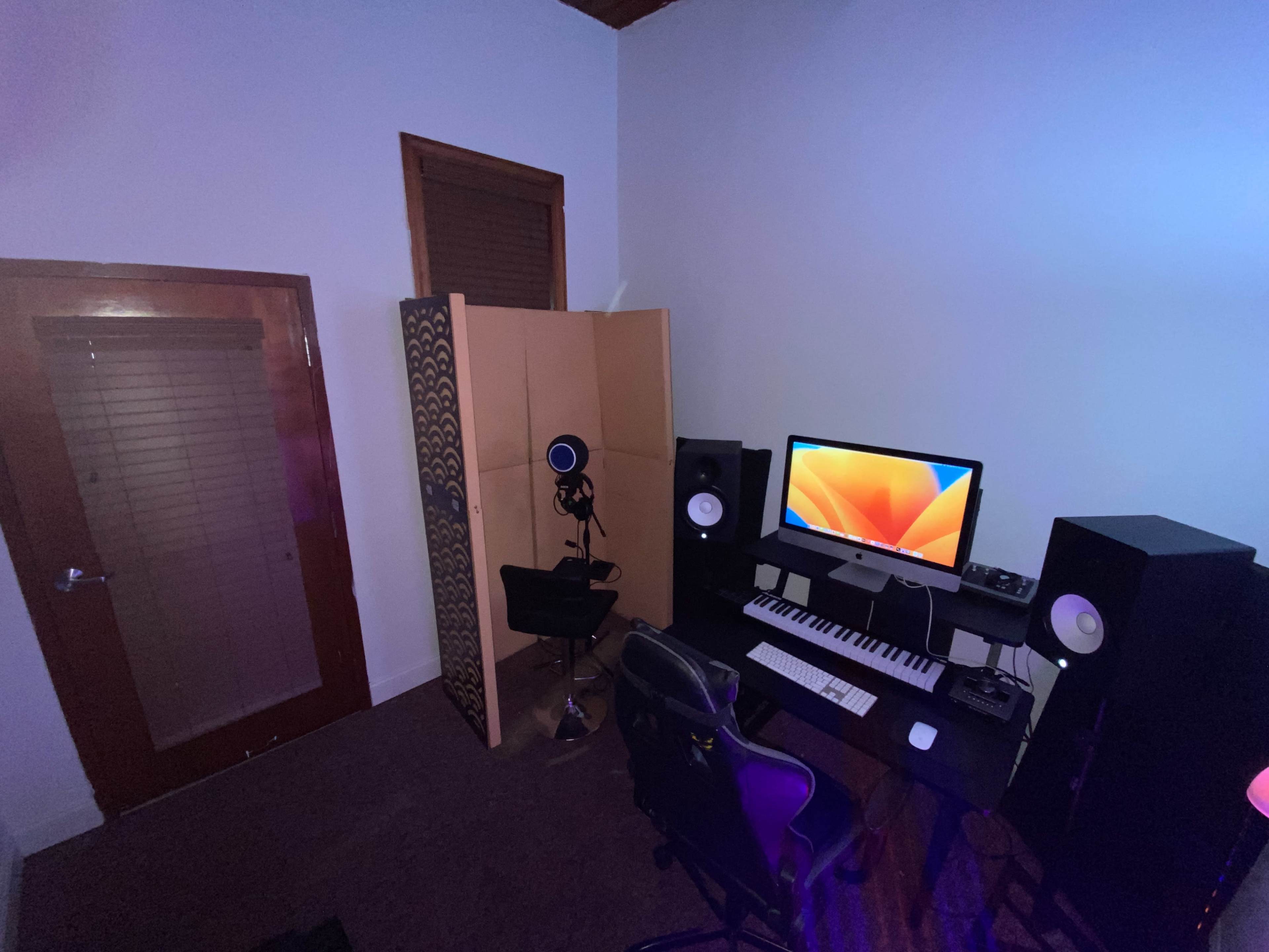The image shows a music production room featuring a computer, keyboard, microphone, speakers, and a privacy screen in a corner setup.