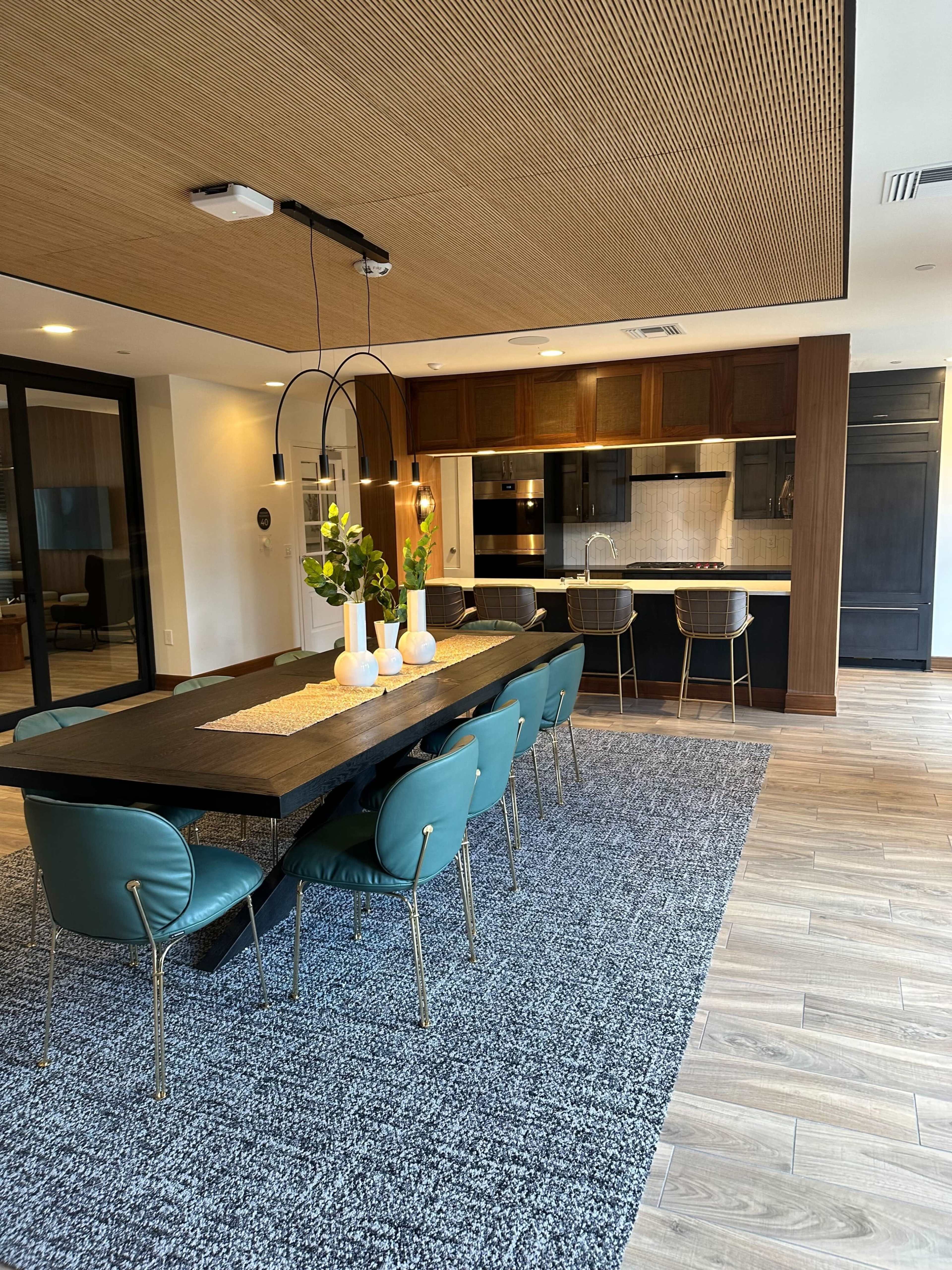 A modern dining area with a large rectangular table, teal chairs, and a view of a kitchen with dark cabinetry and stylish appliances.
