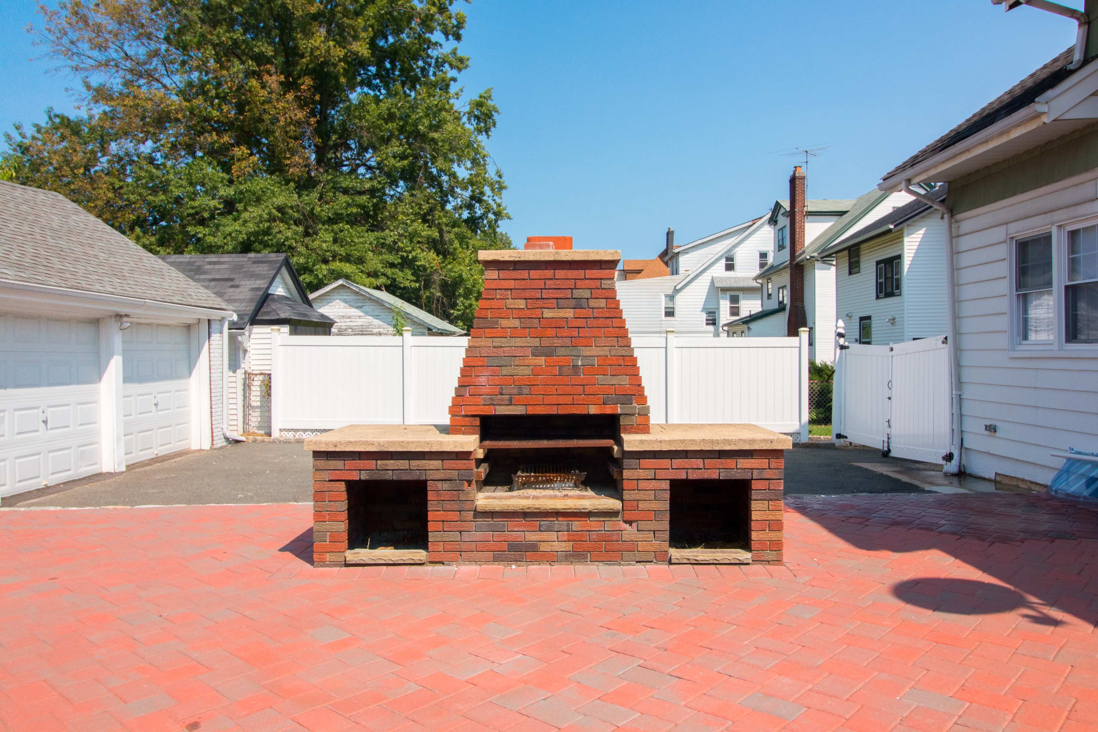 A brick outdoor barbecue grill situated on a paved area, surrounded by residential buildings and a white fence.