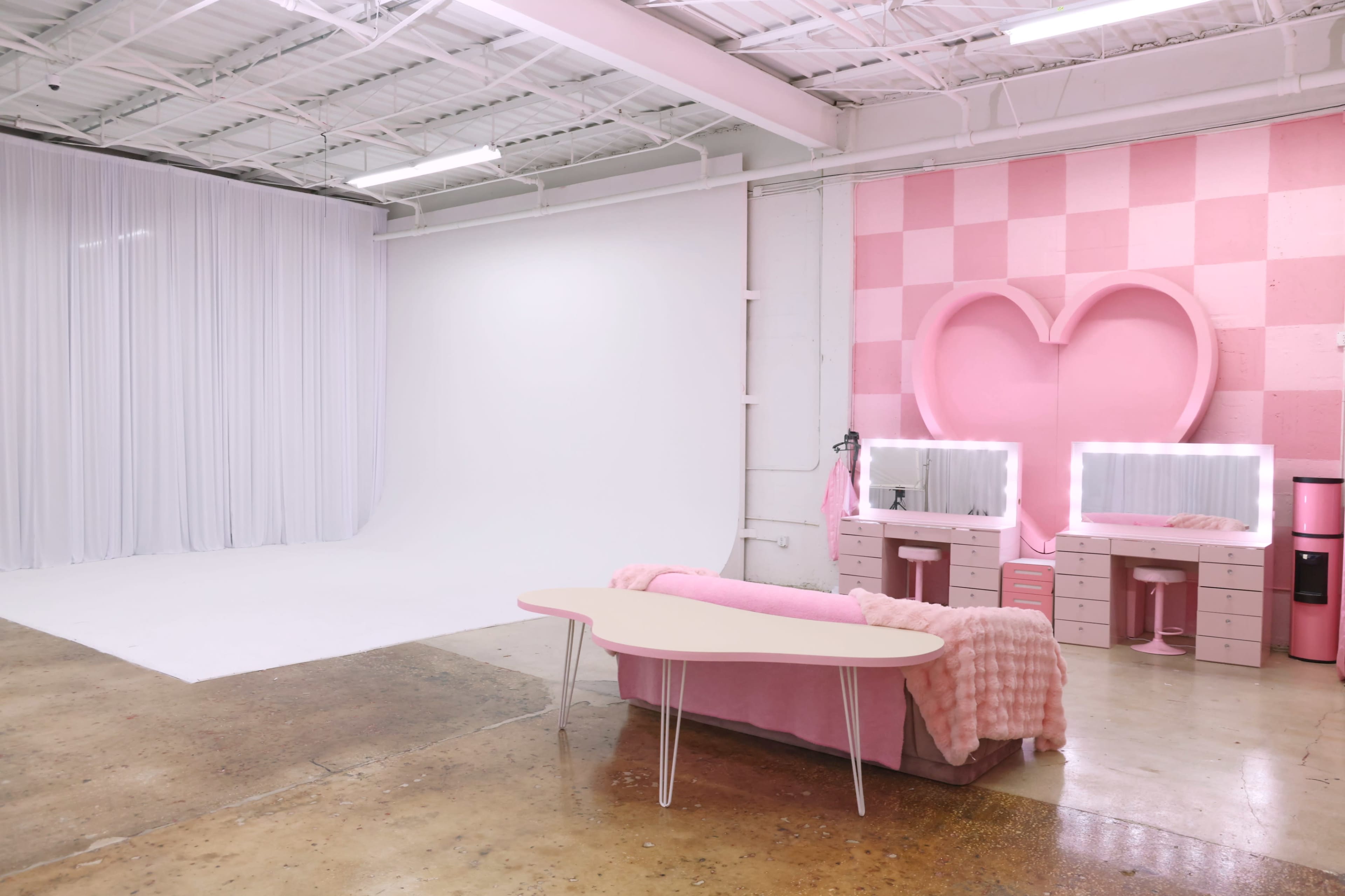 The image shows a bright studio space featuring a white backdrop, a pink heart-shaped wall decor, and a pink couch with a matching blanket.