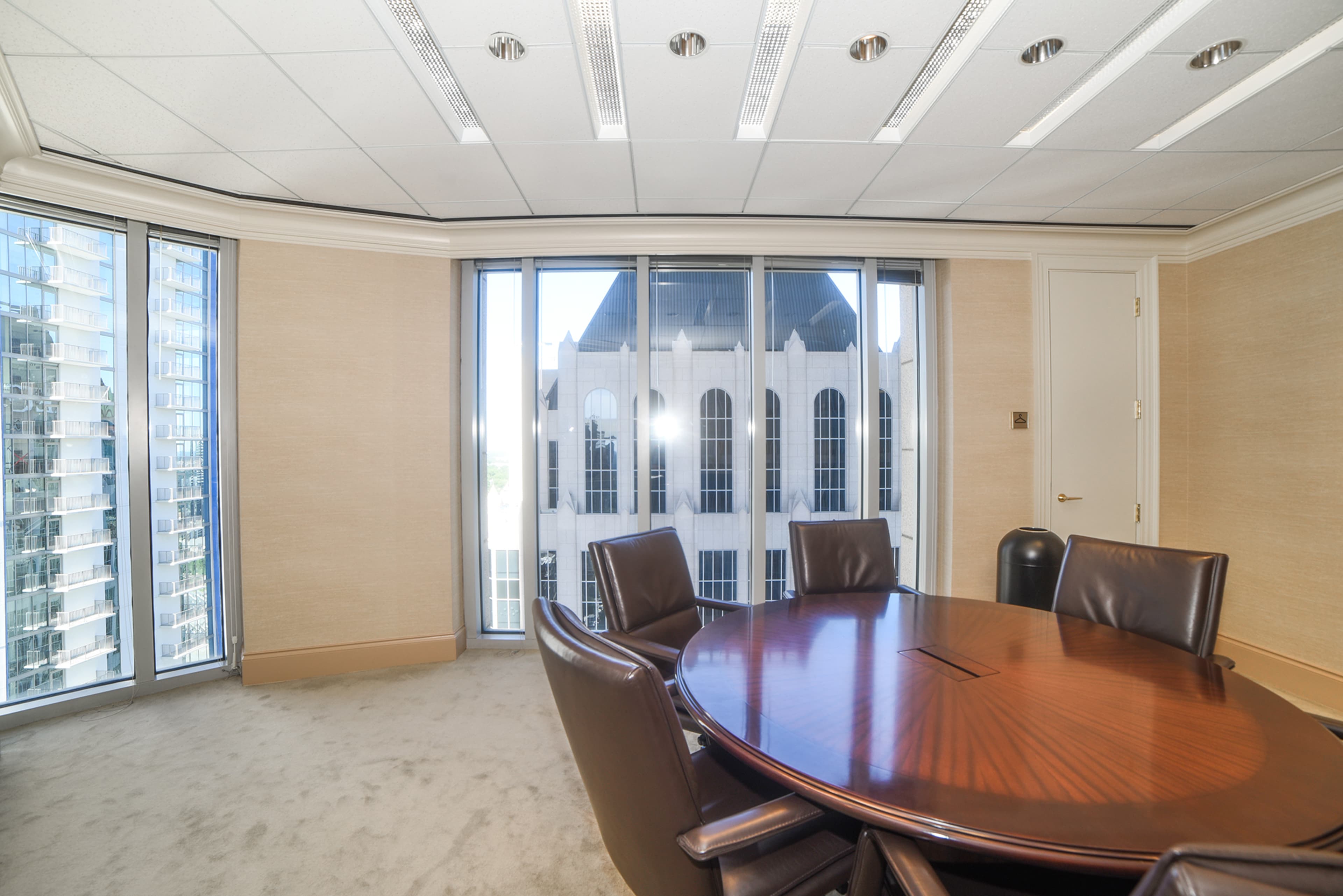 A modern conference room features a large wooden table surrounded by leather chairs, with floor-to-ceiling windows revealing a view of a nearby building.