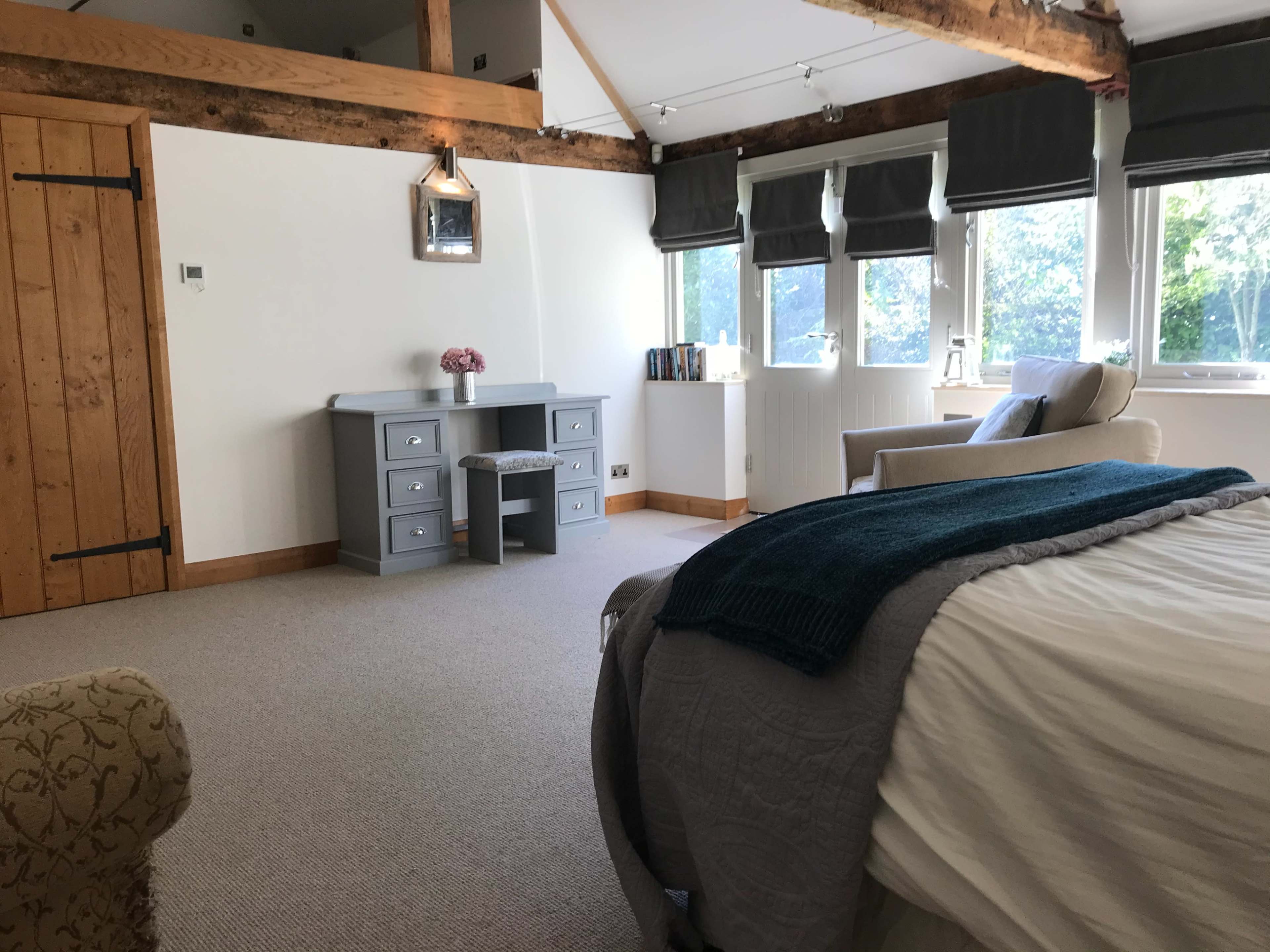 A spacious room with a bed, a desk with drawers, and a chair, featuring large windows and wooden beams.