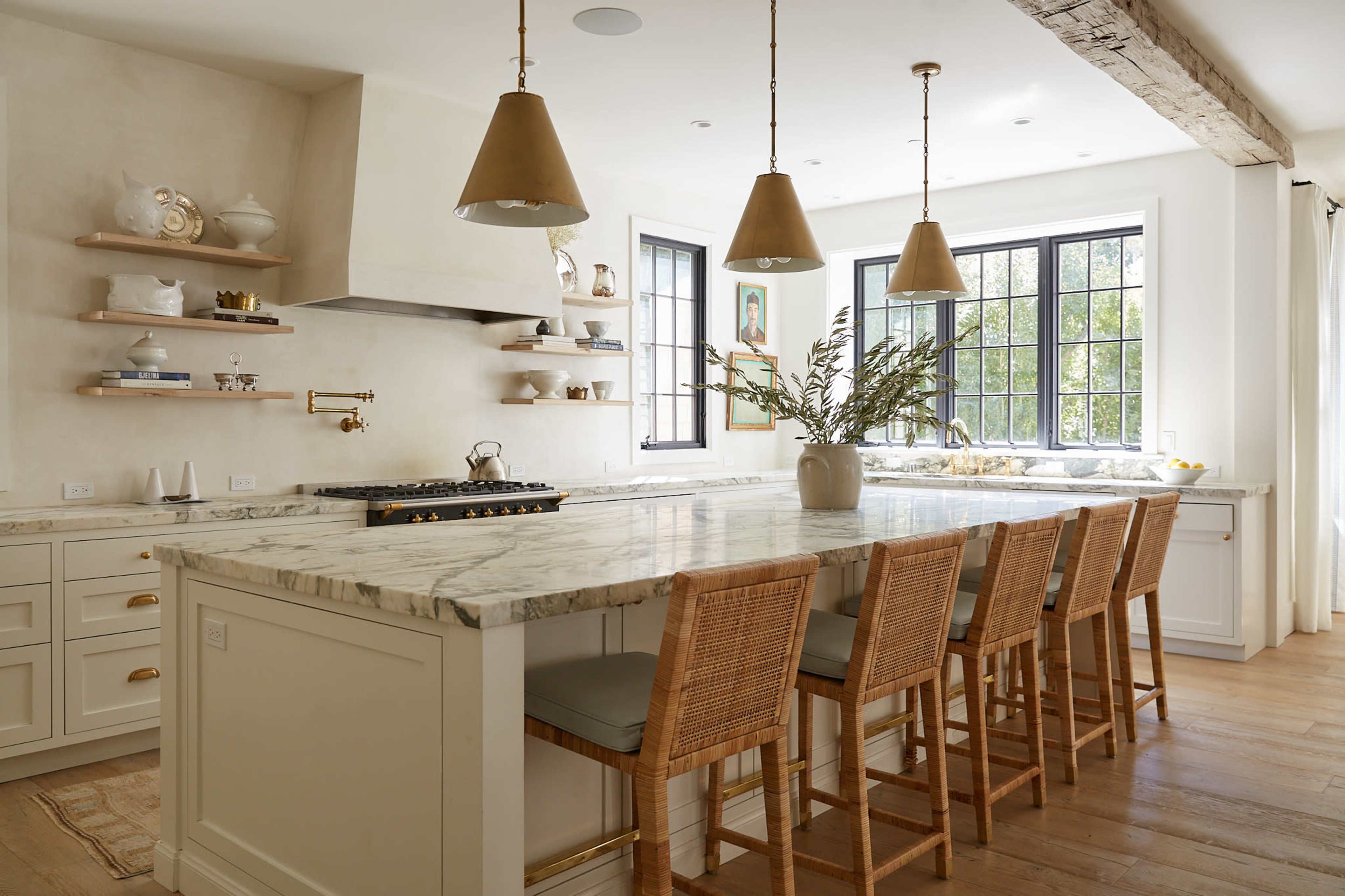 A modern kitchen features a large marble island with bar stools, brass pendant lights, and windows providing natural light.