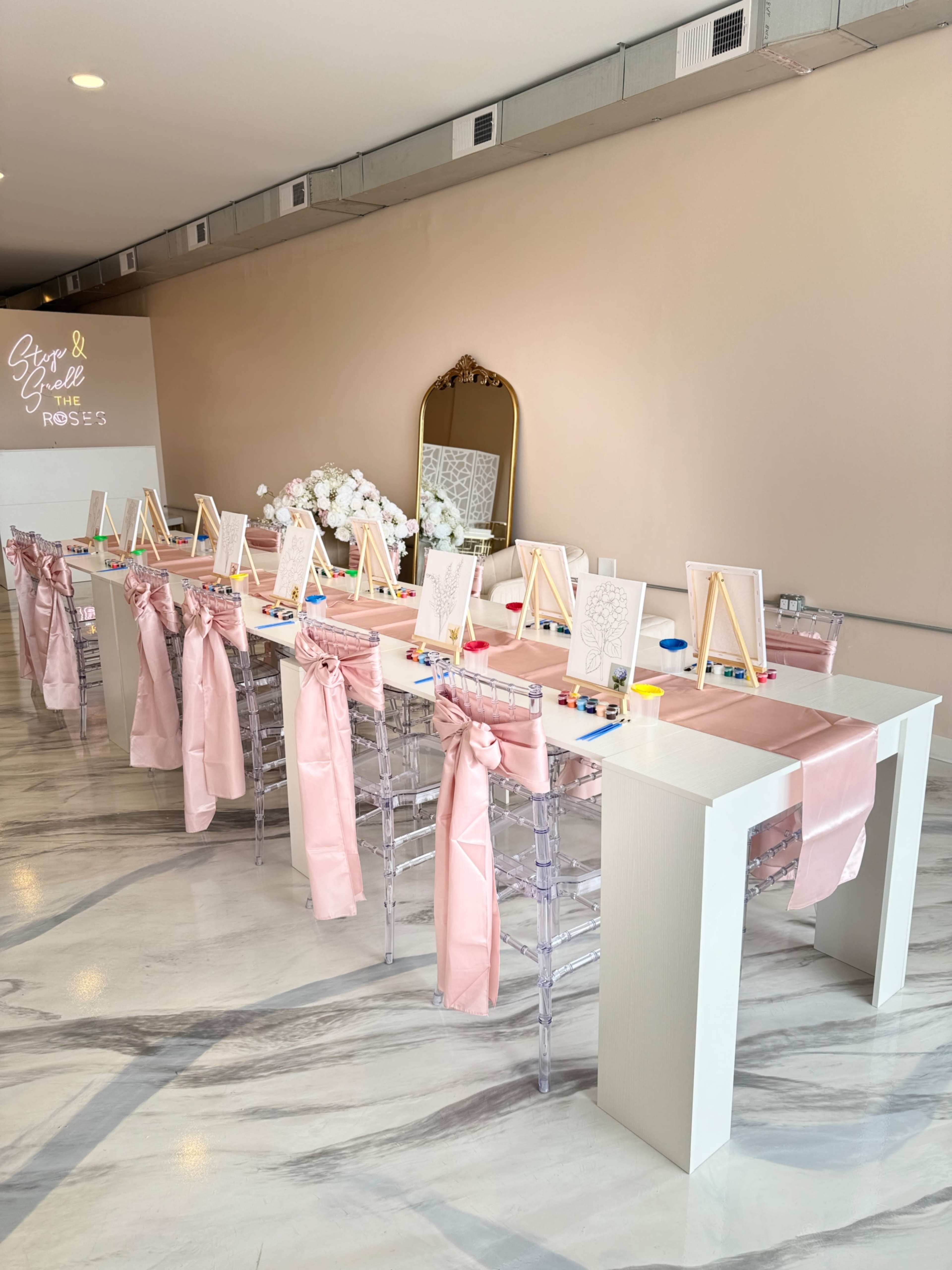 A long table is set for an art activity, featuring pink sashes on clear chairs, small easels, and art supplies arranged along the surface.