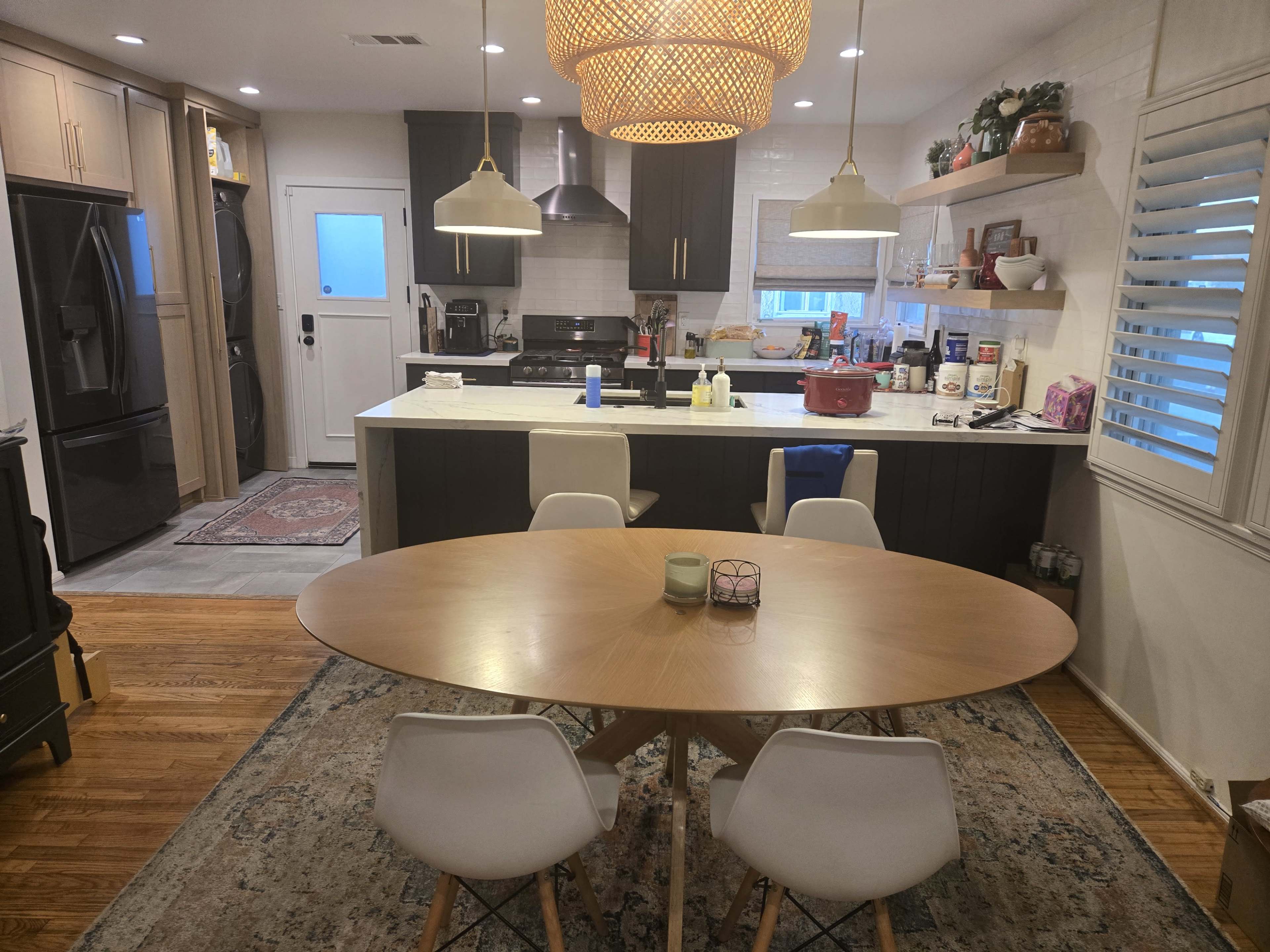 A modern kitchen and dining area features a round wooden table surrounded by four chairs, with a kitchen countertop visible in the background, adorned with various utensils and appliances.