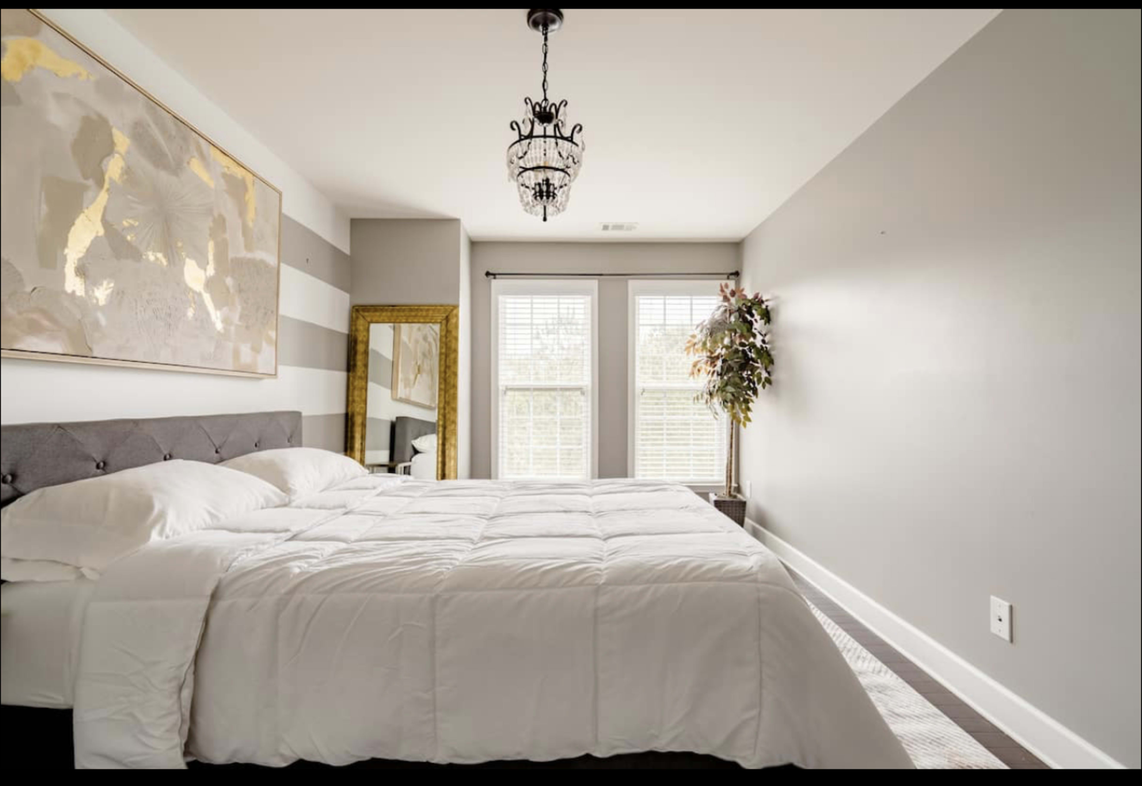 A bedroom features a large bed with white bedding, a striped wall, and windows that allow natural light to enter.
