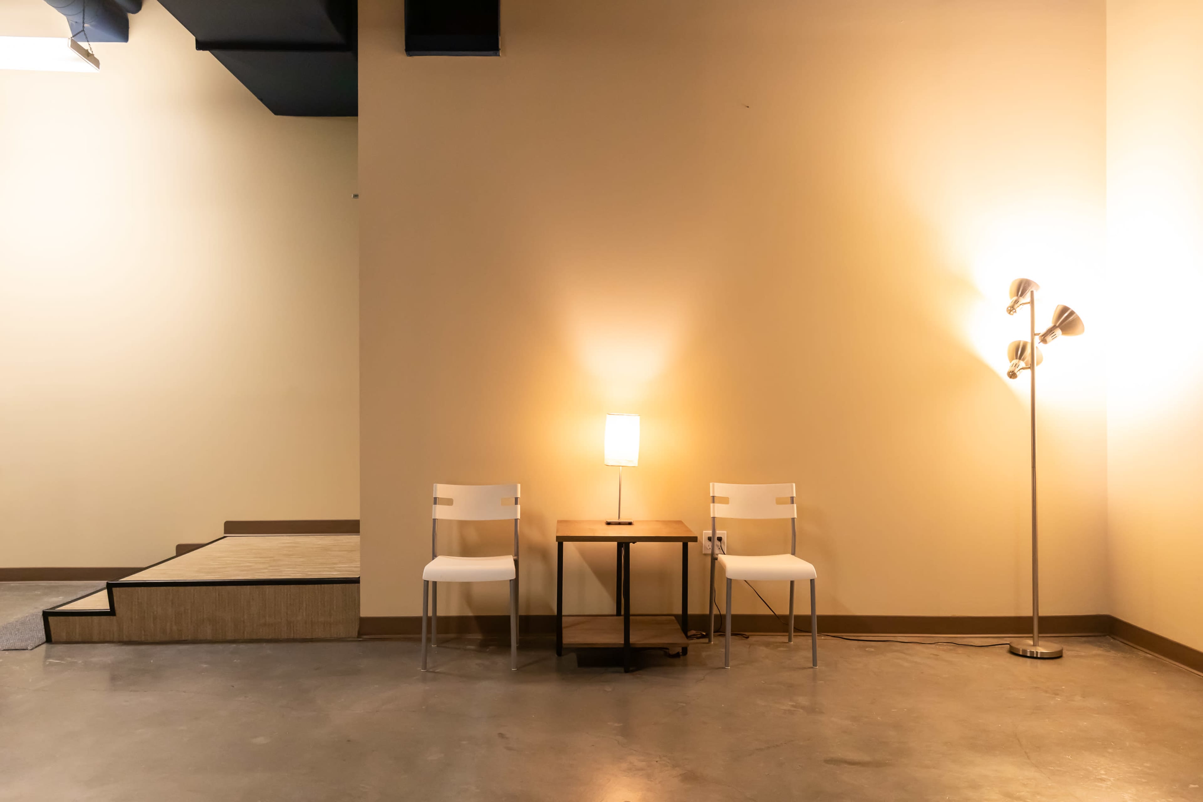 Two white chairs next to a small table with a lamp in a sparsely furnished room featuring a light-colored wall and a staircase in the background.