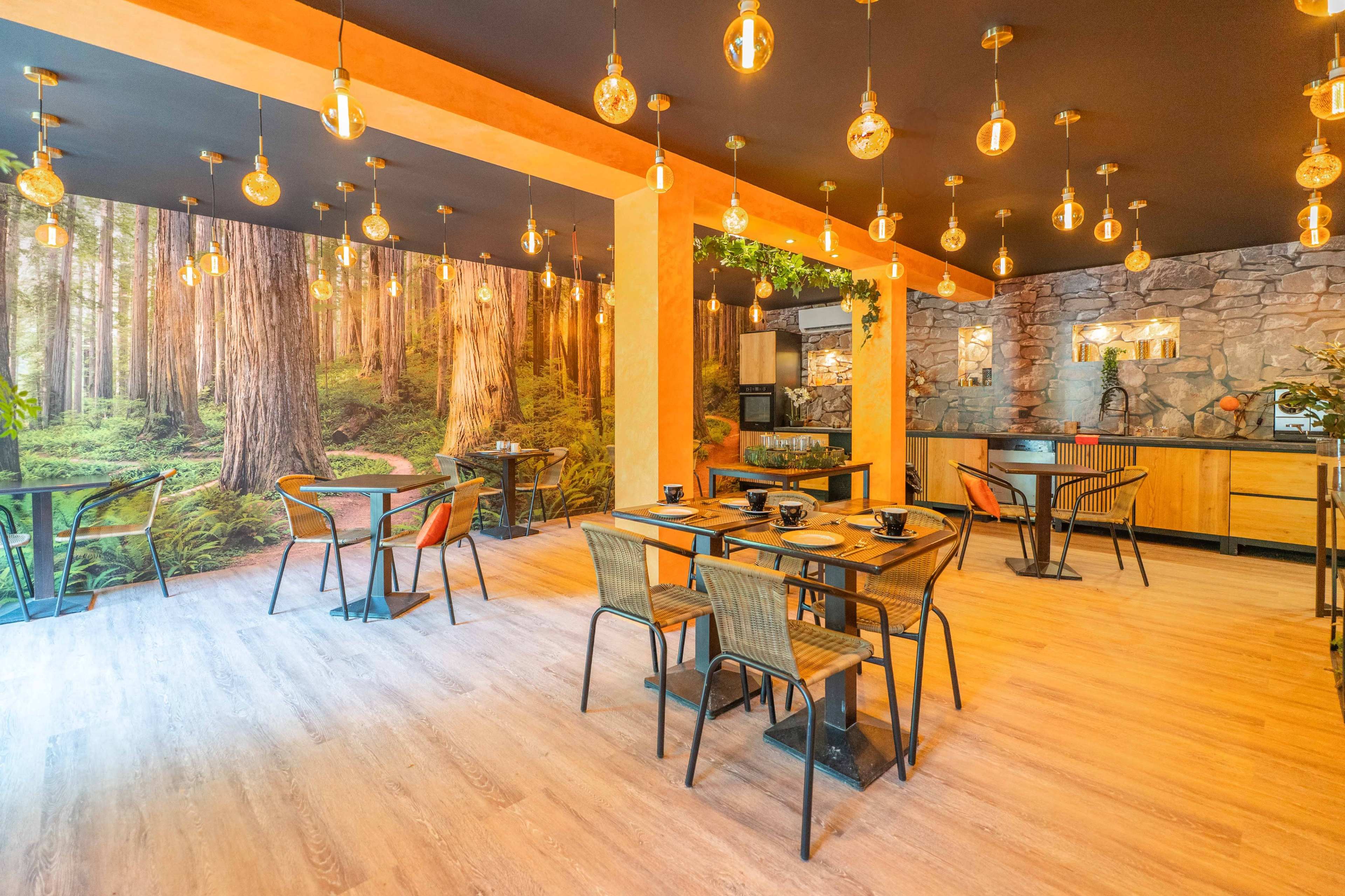The image shows a modern café interior featuring wooden accents, black and metal furniture, and a forest-themed mural on one wall.