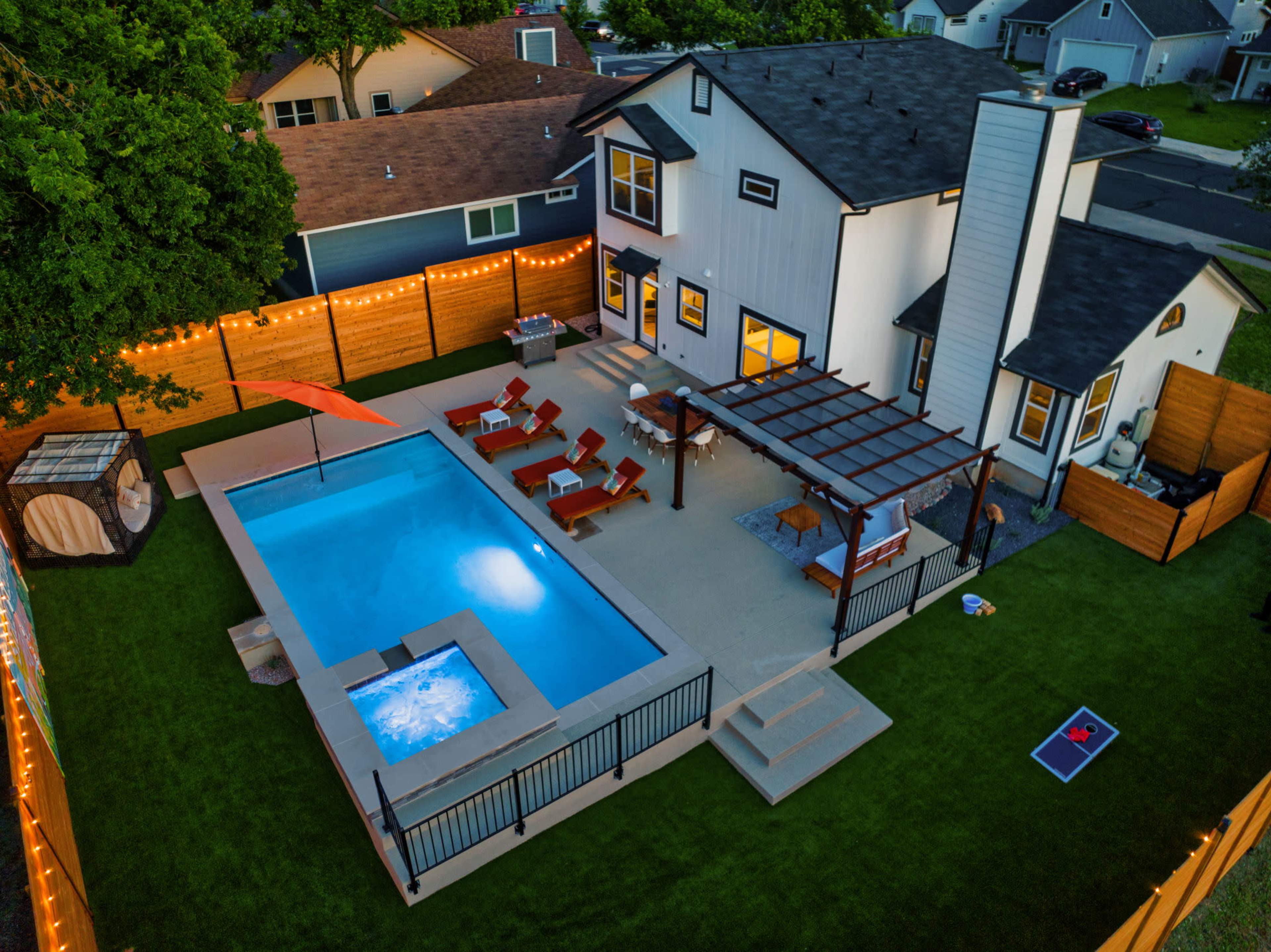 The image shows a backyard featuring a swimming pool, a hot tub, lounge chairs, and a pergola, all surrounded by wooden fencing and string lights.