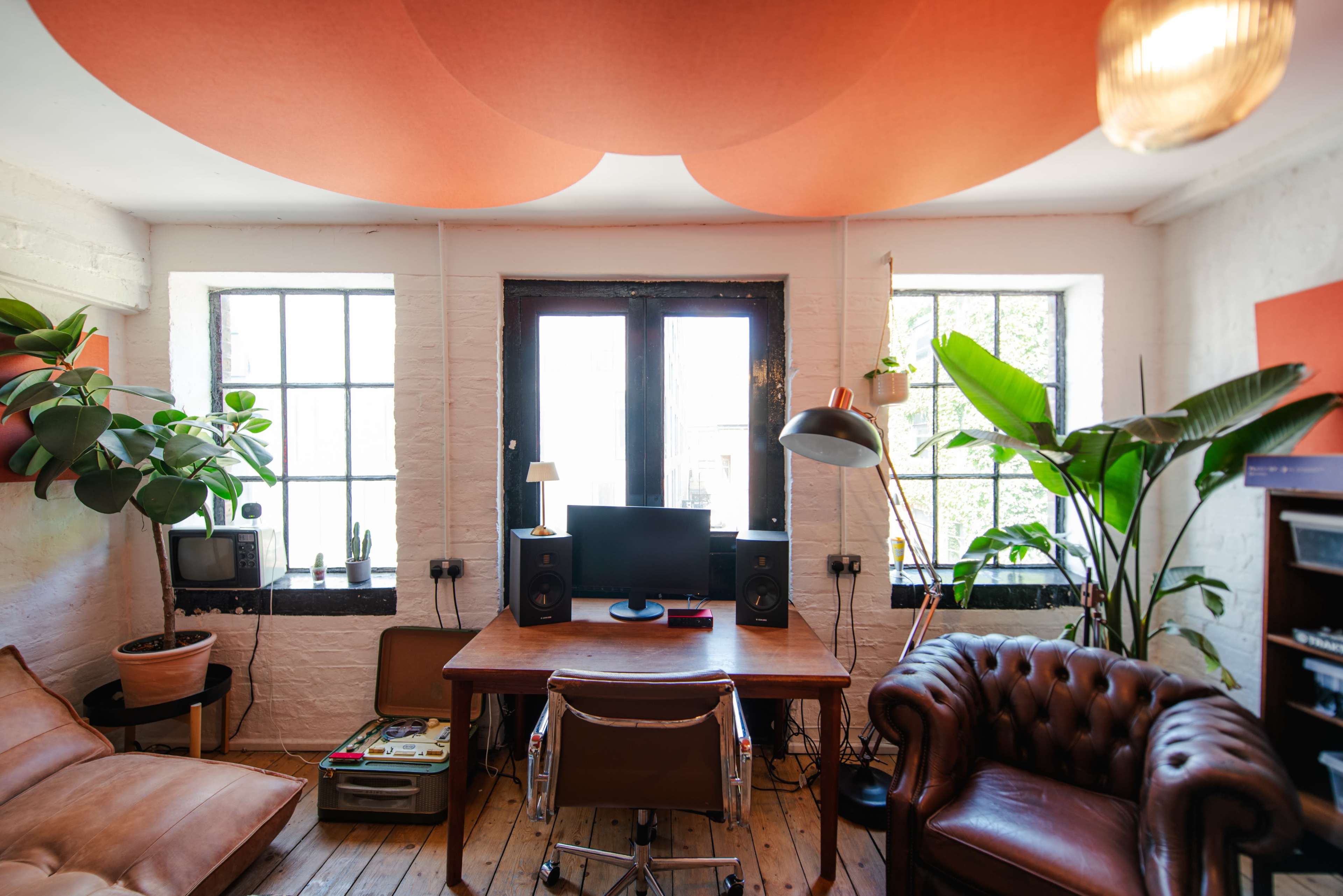 The image shows a cozy office space with large windows, a wooden floor, a desk with a computer, and various plants positioned around the room.