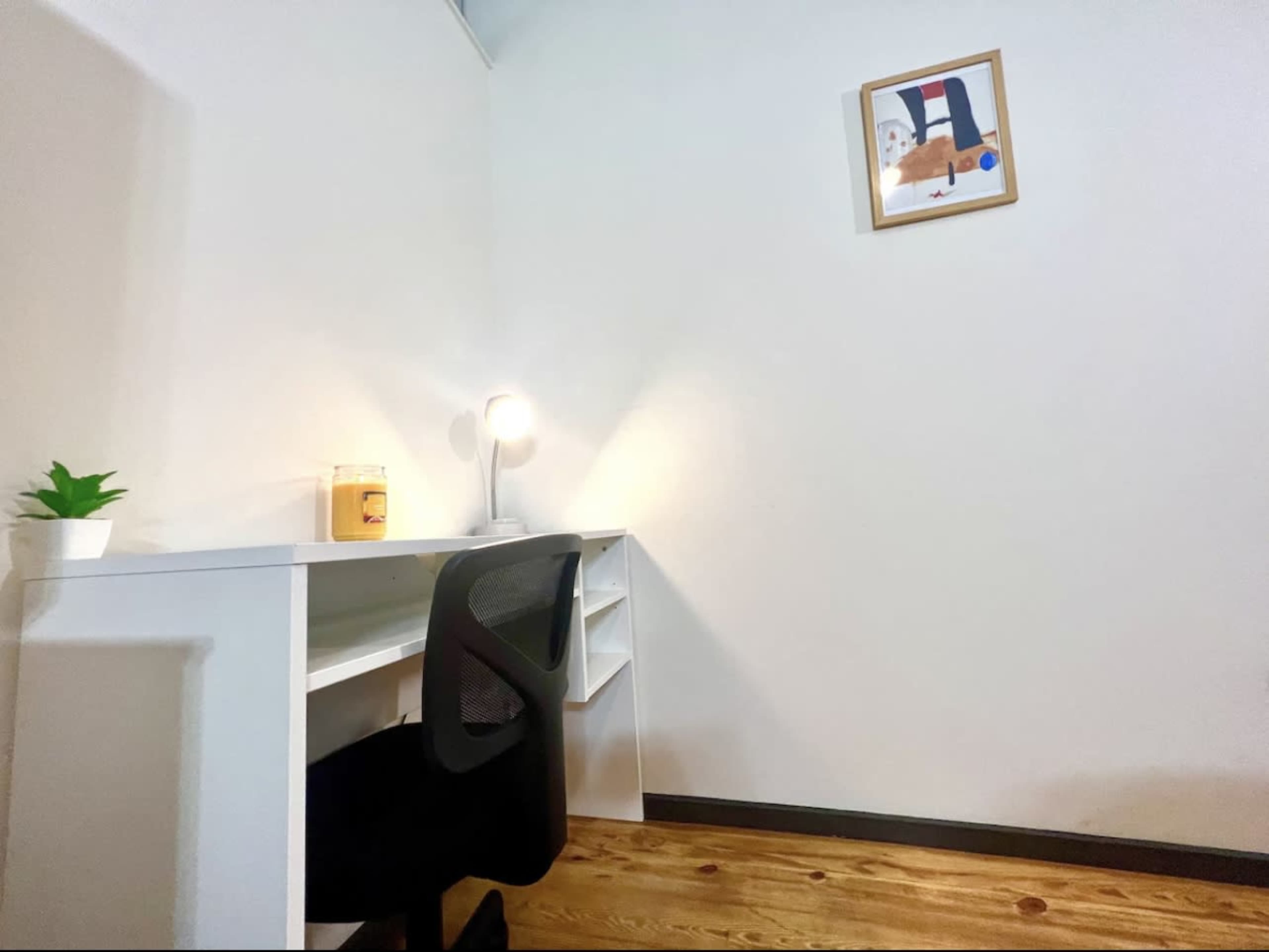 A minimalist desk with a chair, a small potted plant, a lamp, and a framed picture on the wall, set against a neutral-colored wall and wooden floor.