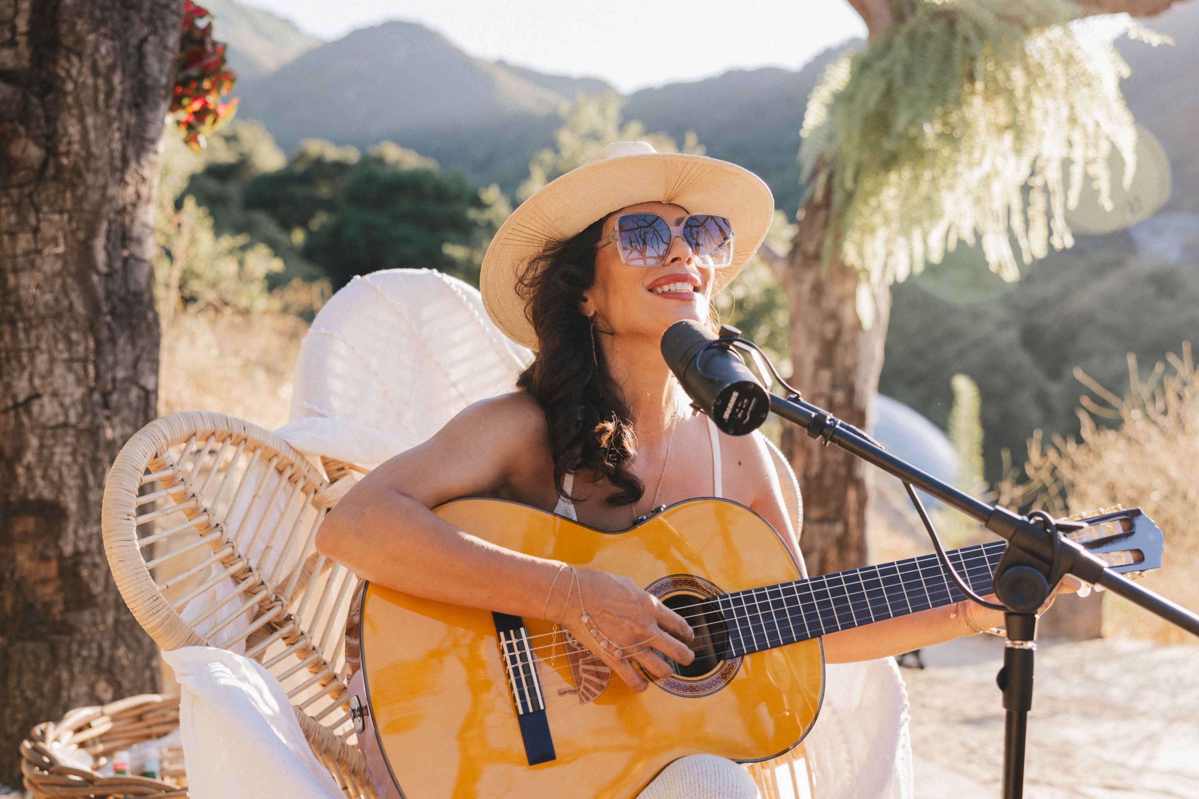 A woman wearing sunglasses and a straw hat sings into a microphone while playing a guitar in a scenic outdoor setting.