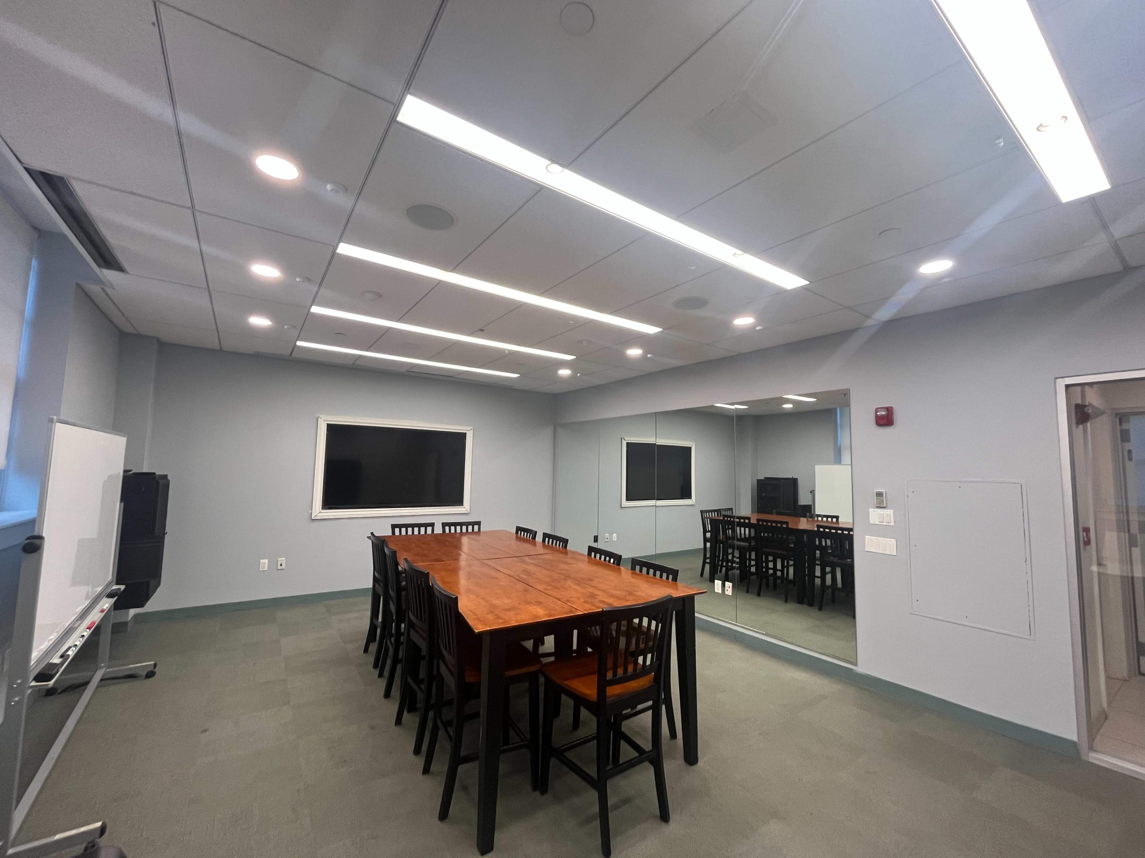 A modern conference room features a large wooden table surrounded by black chairs, with a flat-screen monitor and large windows providing bright overhead lighting.
