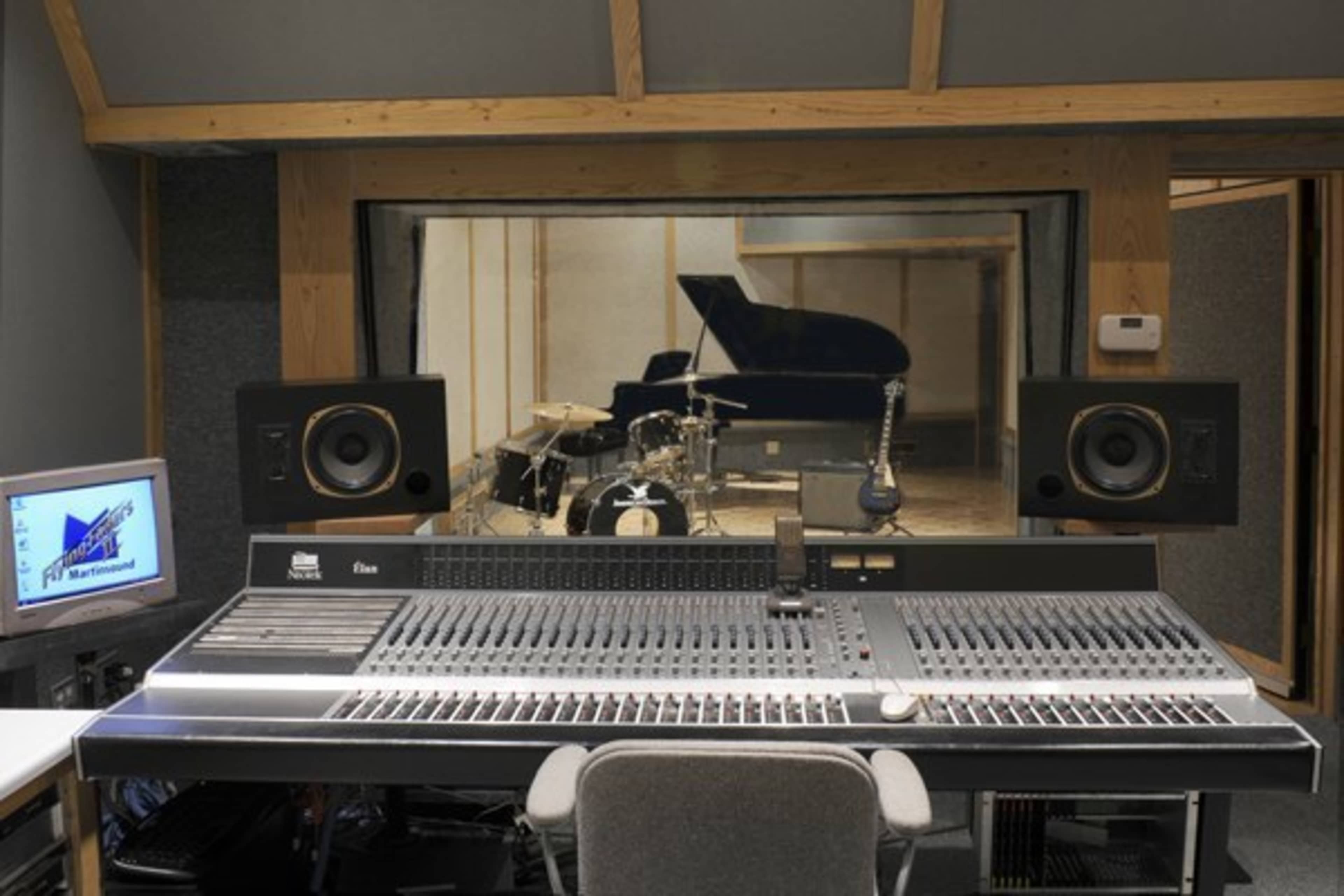 A music recording studio featuring a large mixing console, speakers, and a glass-walled room with a piano, guitar, and drums.