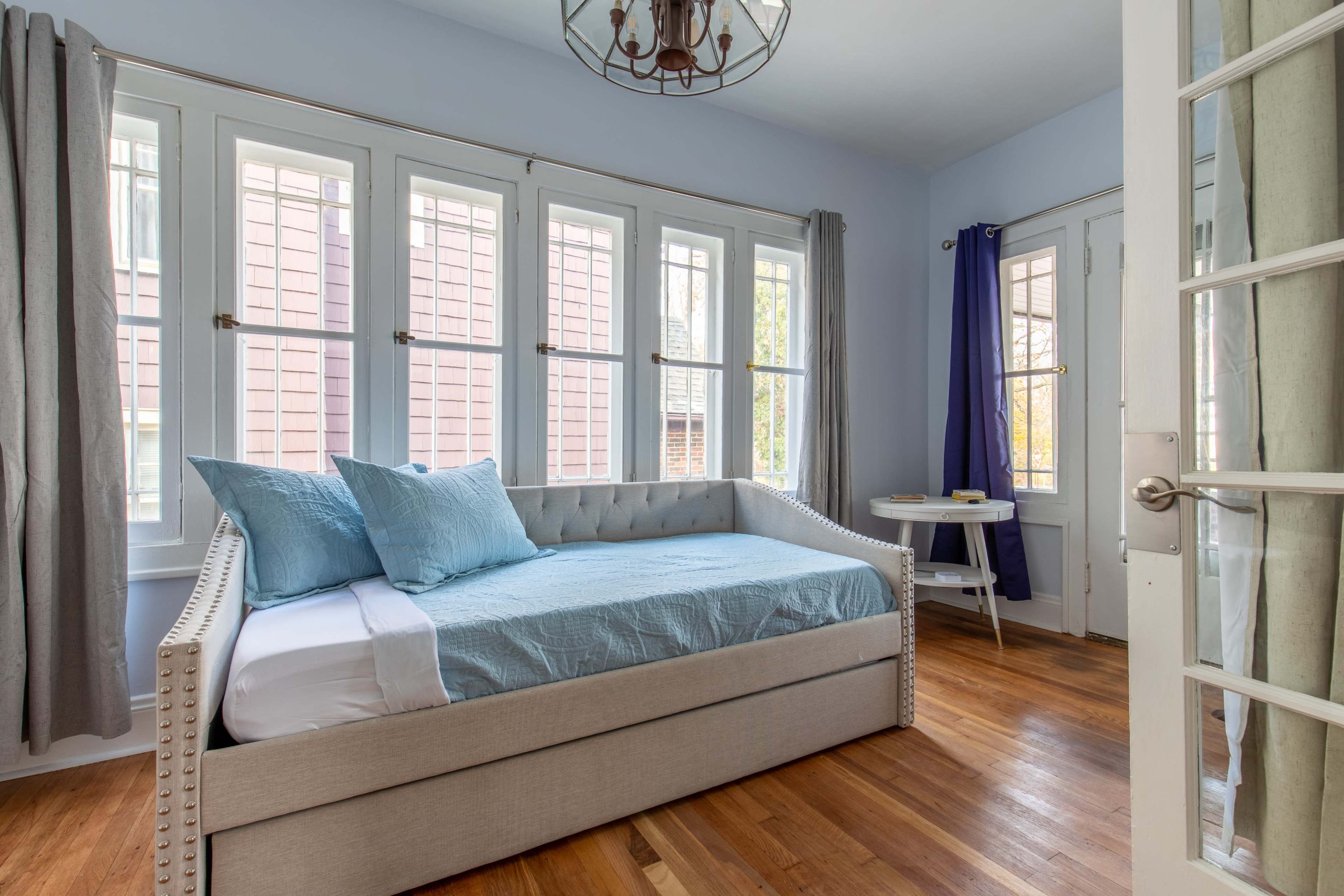 A light blue room features a daybed with blue bedding, large windows, and a small round table nearby.