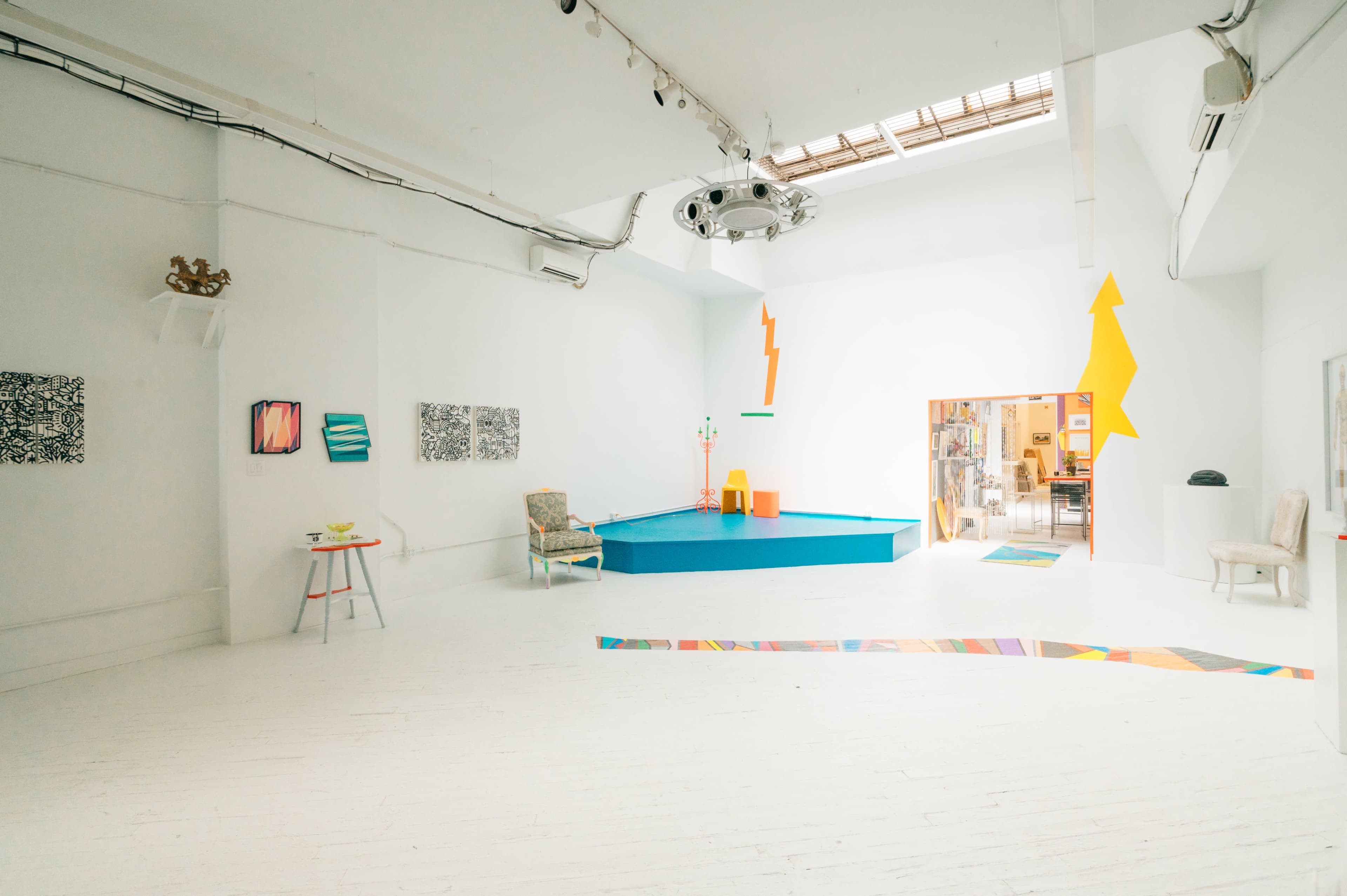 A bright, minimalist art gallery featuring a blue platform in the center, surrounded by colorful artwork on the white walls.