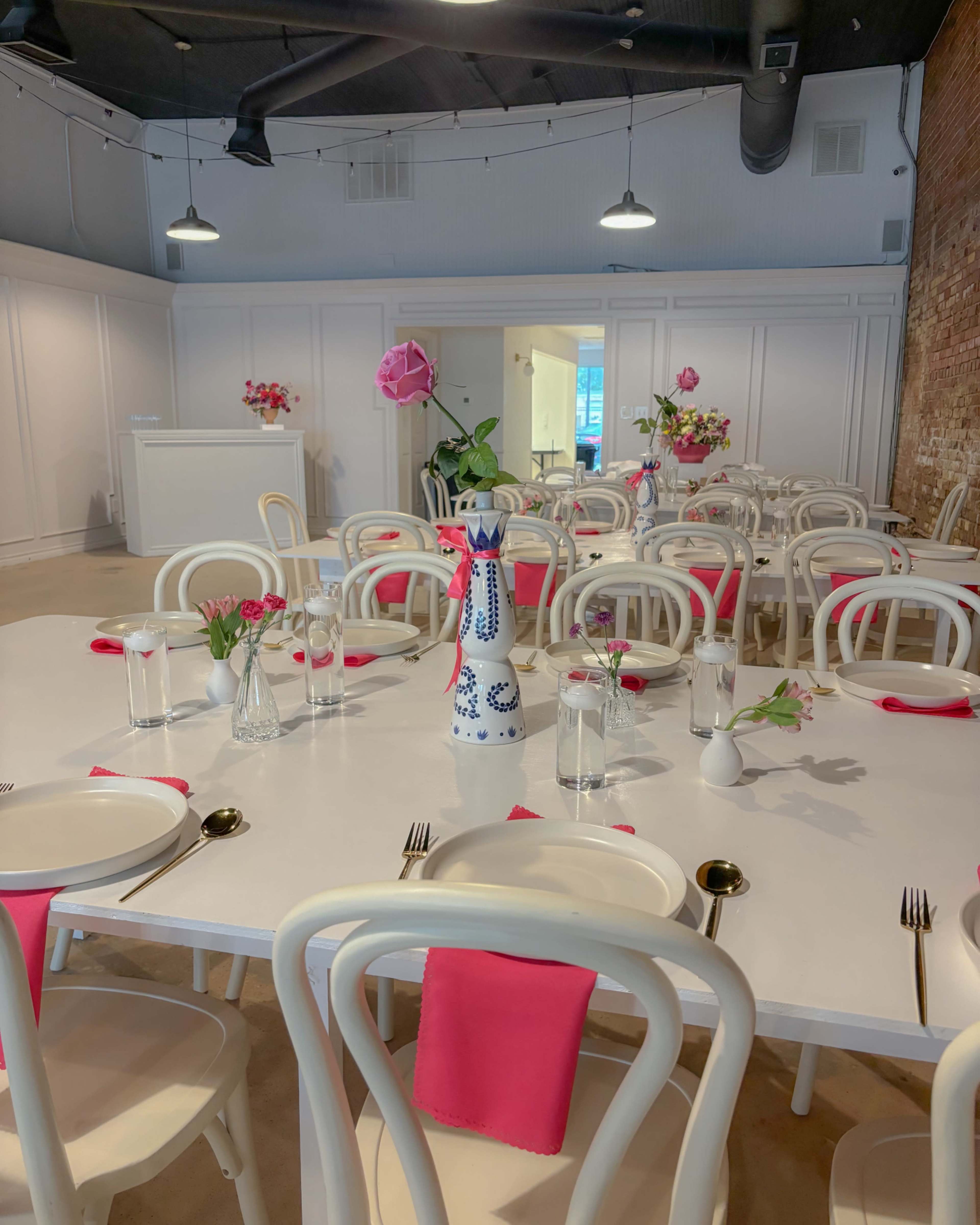 A brightly lit dining area features white tables set with pink napkins, glass vases with flowers, and white chairs.