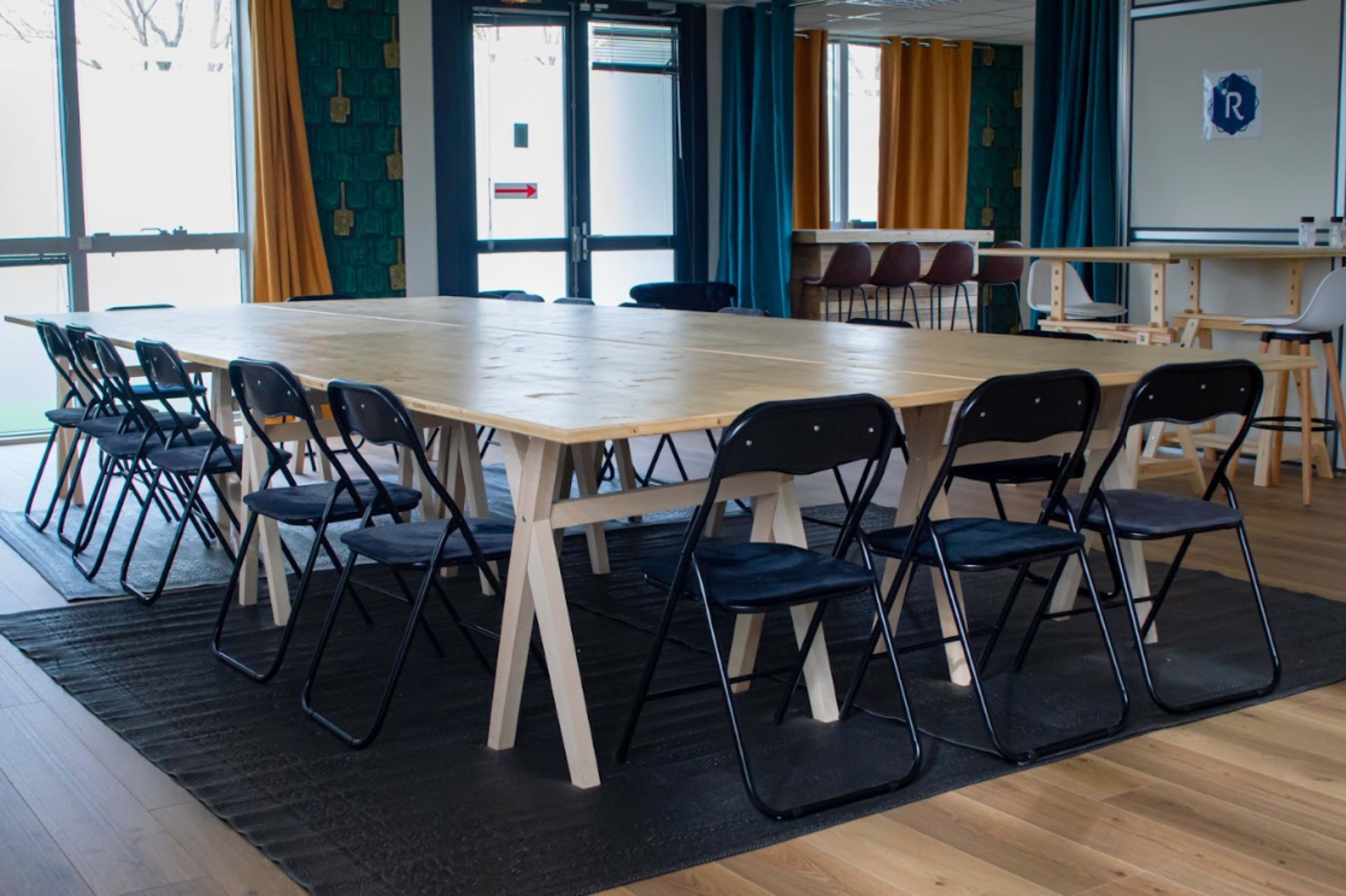 A large, rectangular wooden table with black folding chairs is set up in a brightly lit meeting room featuring large windows and colorful curtains.