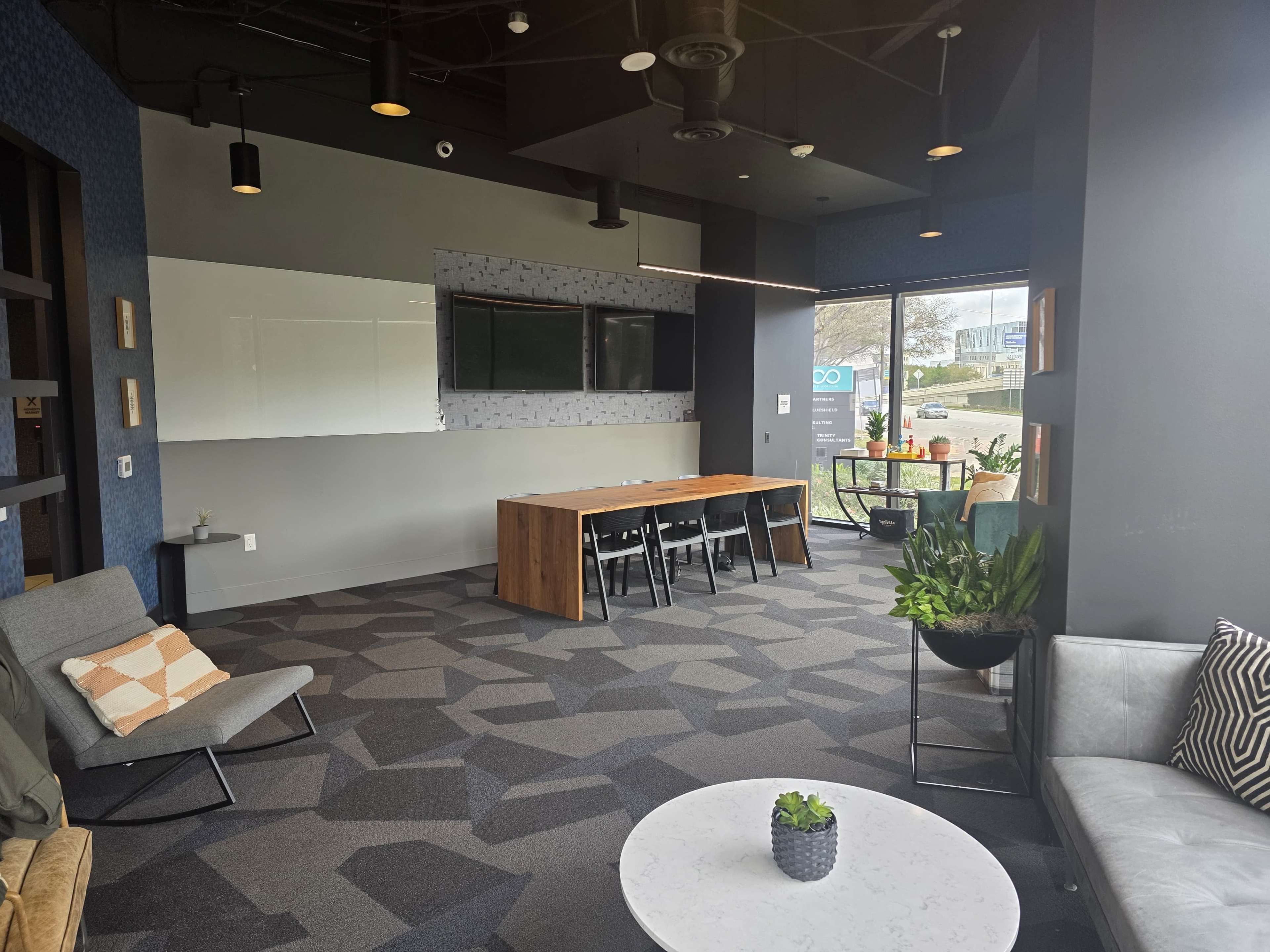 The image shows a modern office space featuring a large table with chairs, a lounge area with seating, and plants, all set against a backdrop of large windows.