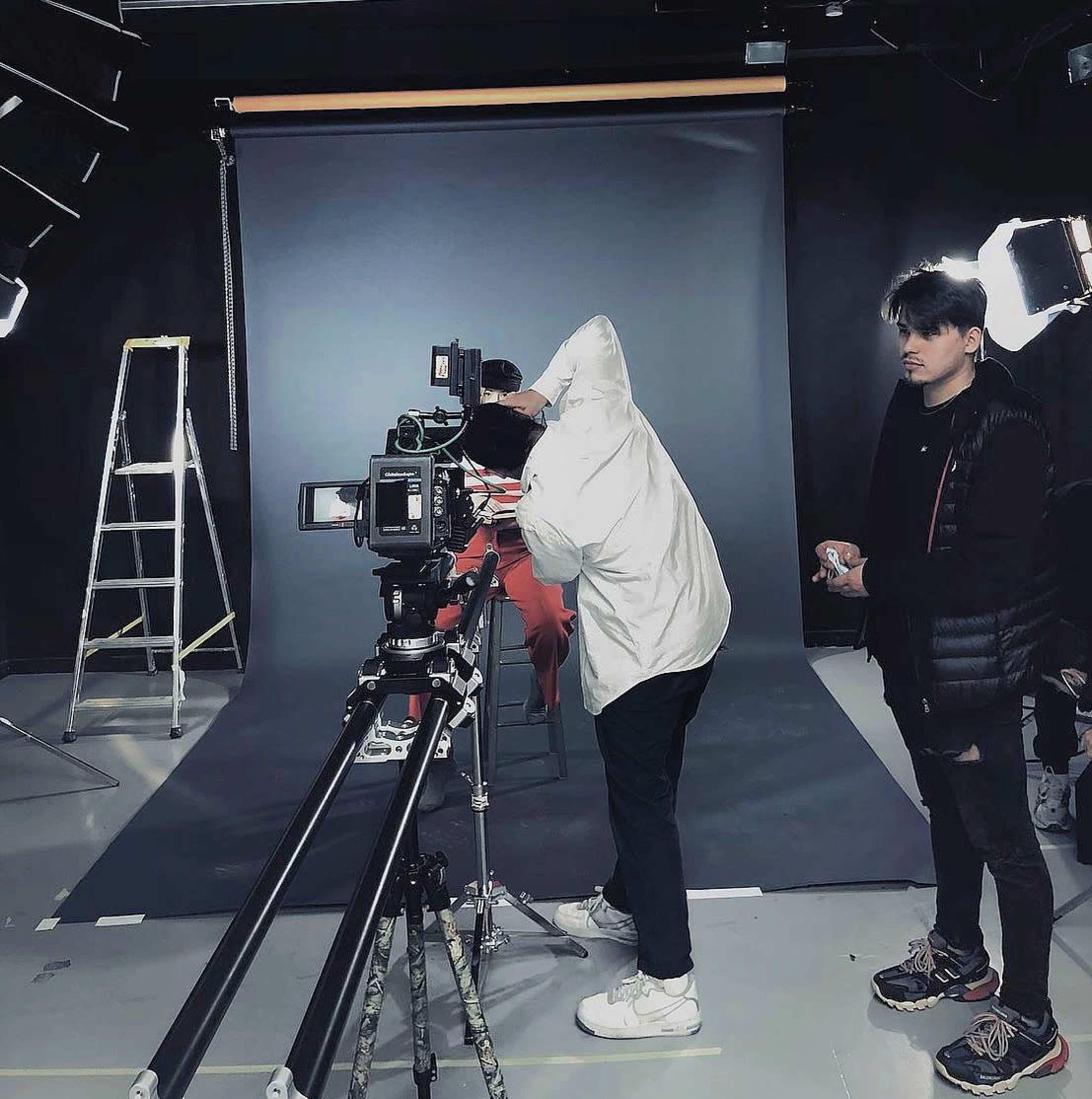 A person in a white jacket operates a camera on a tracking dolly while another individual stands nearby in a dimly lit studio with a backdrop and lighting equipment.