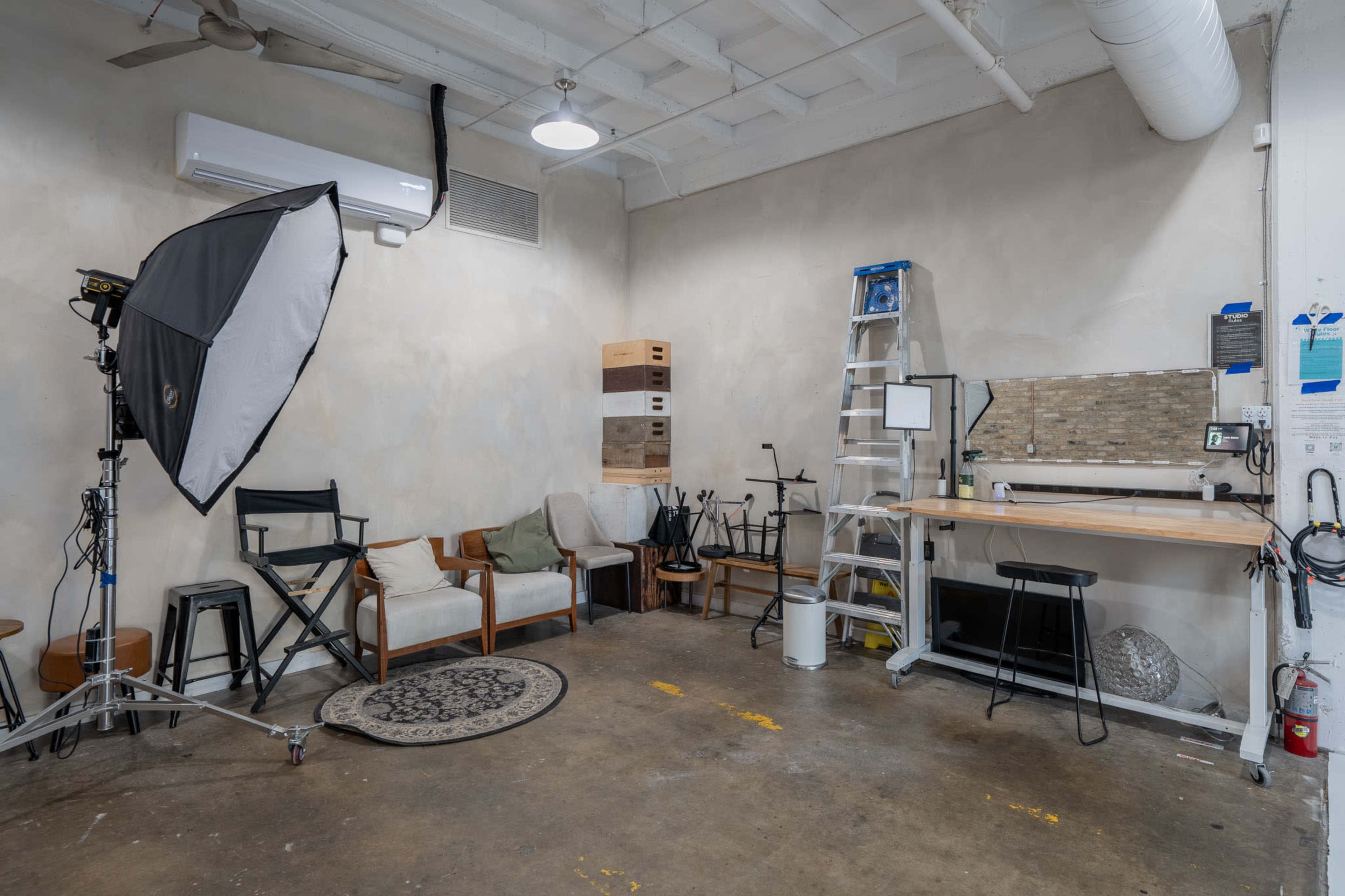 The image shows a photography studio with a backdrop, seating area, a ladder, and equipment storage on the walls.