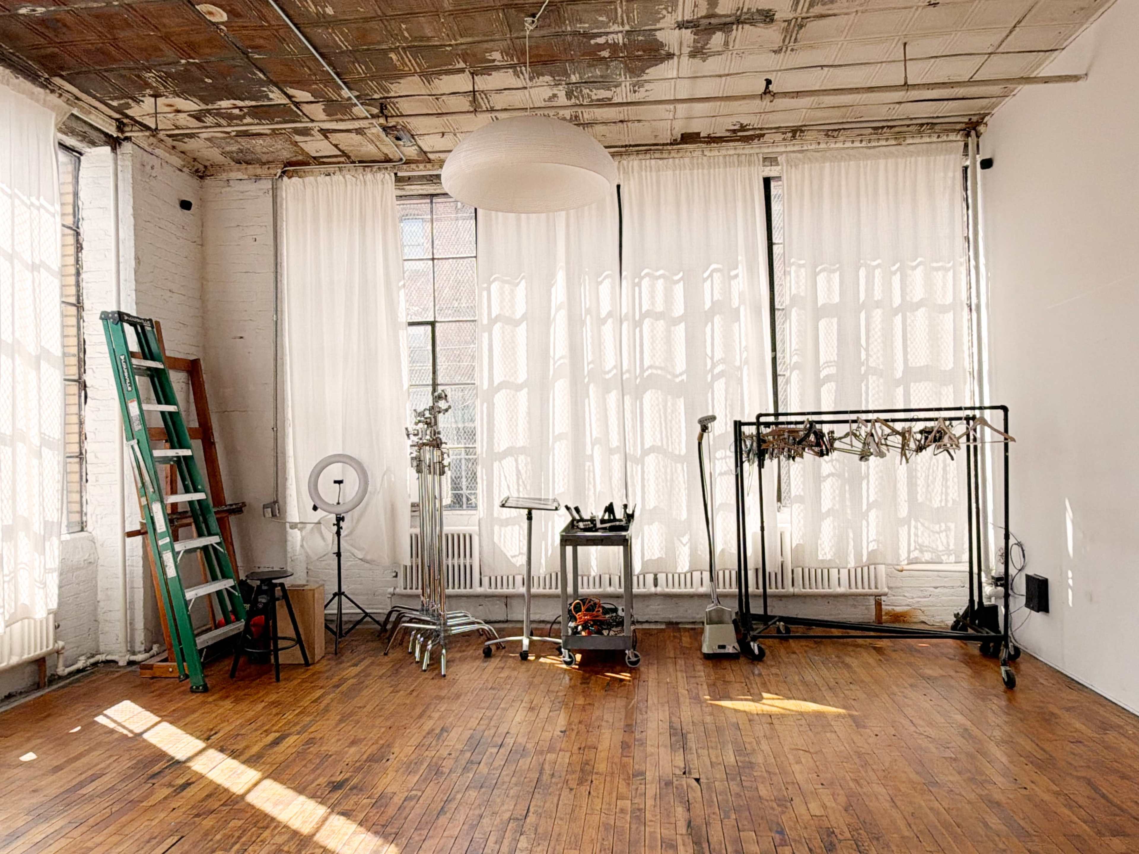 The image shows an empty industrial-style room with large windows, a wooden floor, a green ladder, and a clothing rack with various items hanging on it.