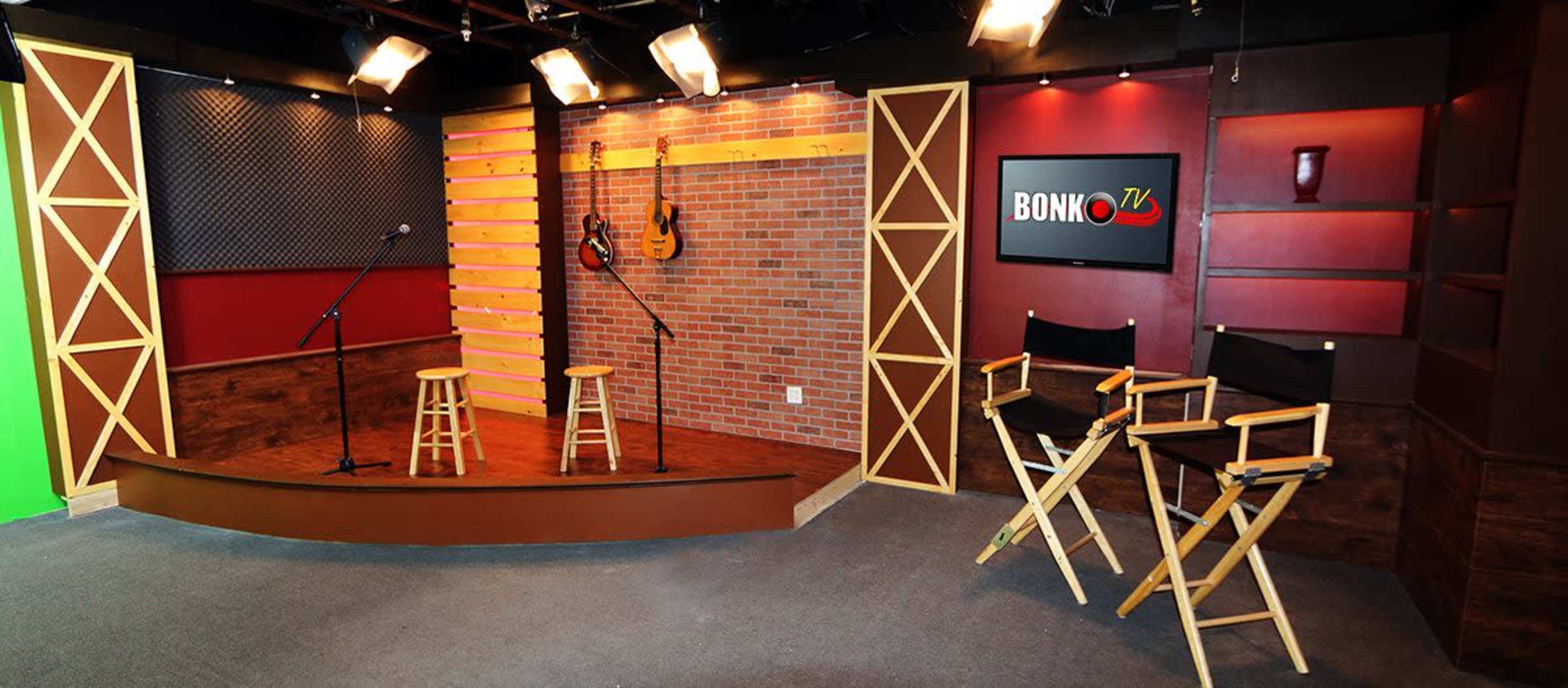 The image shows a television studio set featuring two microphone stands, two stools, and director's chairs against a brick backdrop with decorative lighting and guitars on display.